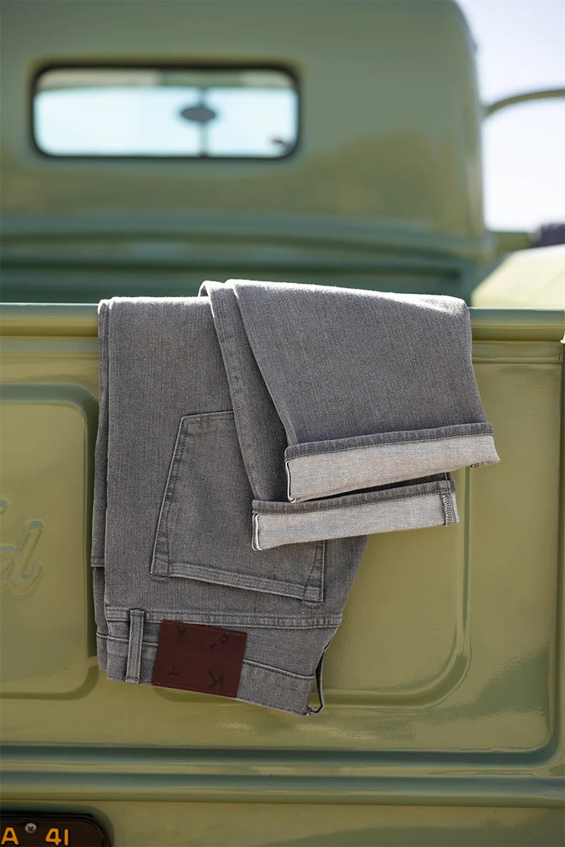 A pair of Hiroshi Kato - The Hammer Straight / Granite 10.5oz Selvedge jeans, featuring a visible brown patch and made from premium Japanese selvedge denim, is draped over the tailgate of a light green vintage truck on a sunny day.