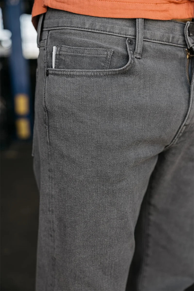 A close-up of a person wearing the Hiroshi Kato - The Hammer Straight / Granite 10.5oz Selvedge jeans and an orange shirt, highlighting the front pocket and part of the waistband. The background is blurred.