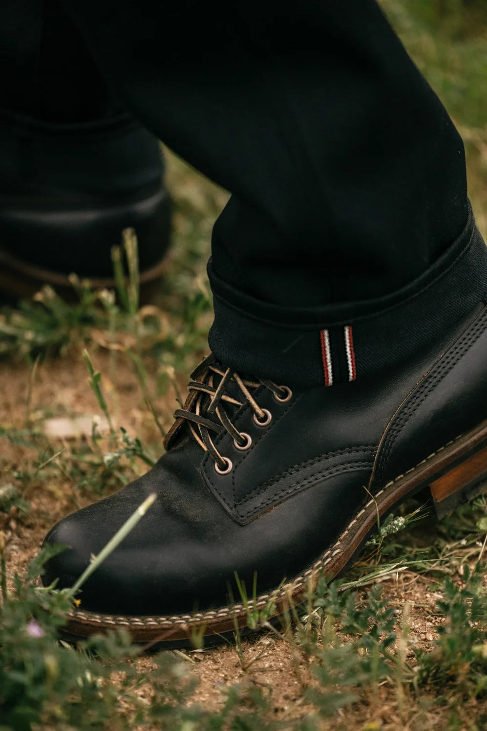 A close-up of a person wearing black leather boots and Hiroshi Kato - The Hammer Straight / Black Raw 14oz Selvedge pants, standing on grassy ground. The cuff of the denim reveals a striped selvedge detail above the boot.