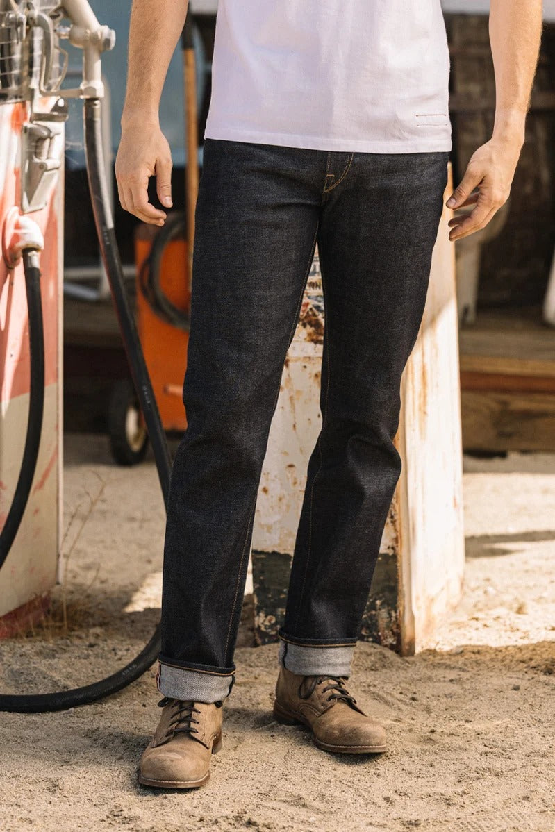 A person in a white shirt, Hiroshi Kato The Hammer Straight / Indigo Raw 14oz Selvedge jeans, and brown boots stands outdoors near an old gas pump on sandy ground.