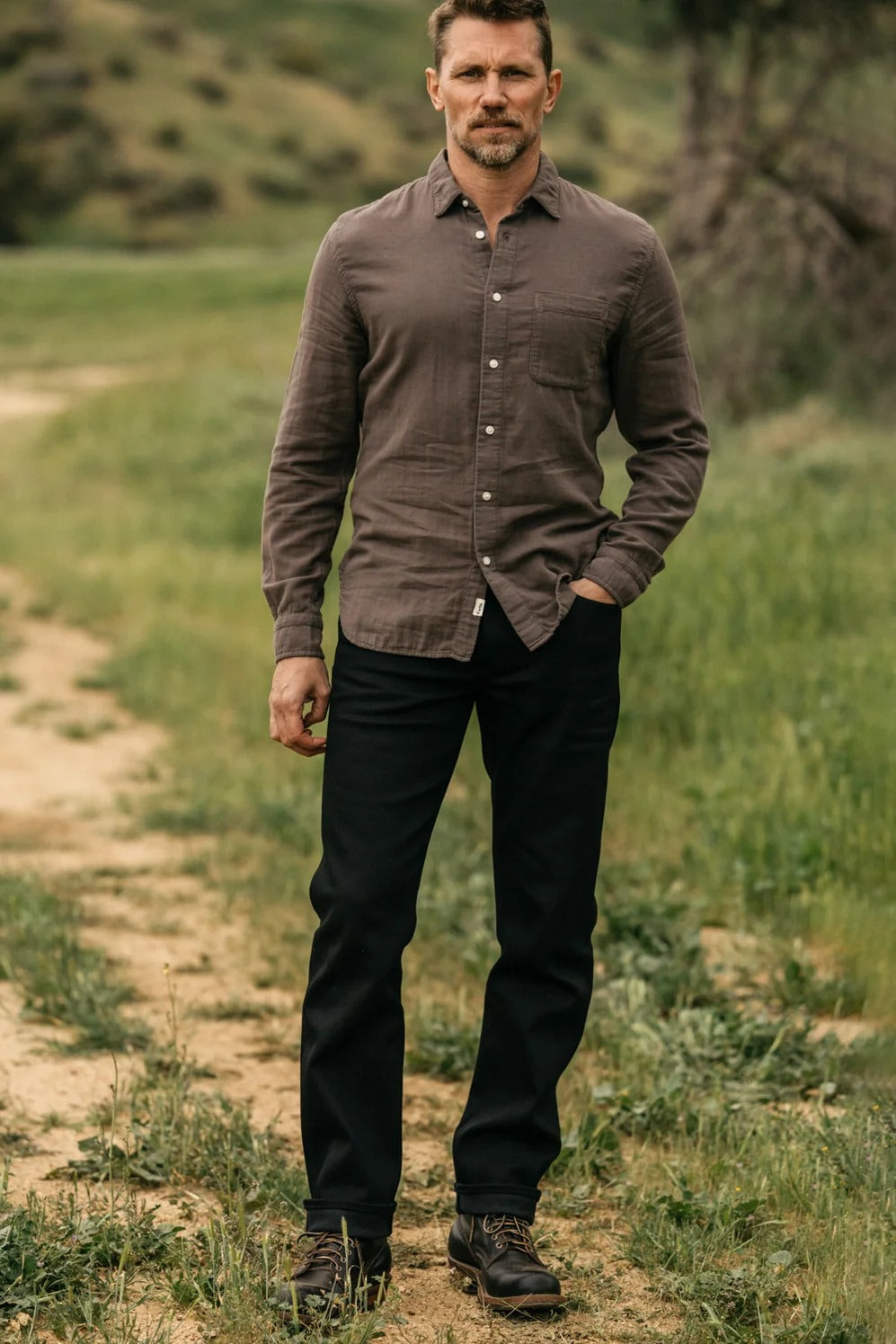 A man stands outdoors on a dirt path, wearing a brown button-up shirt, brown shoes, and Hiroshi Kato - The Hammer Straight / Black Raw 14oz Selvedge jeans by Hiroshi Kato. Green grass and hills are in the background.