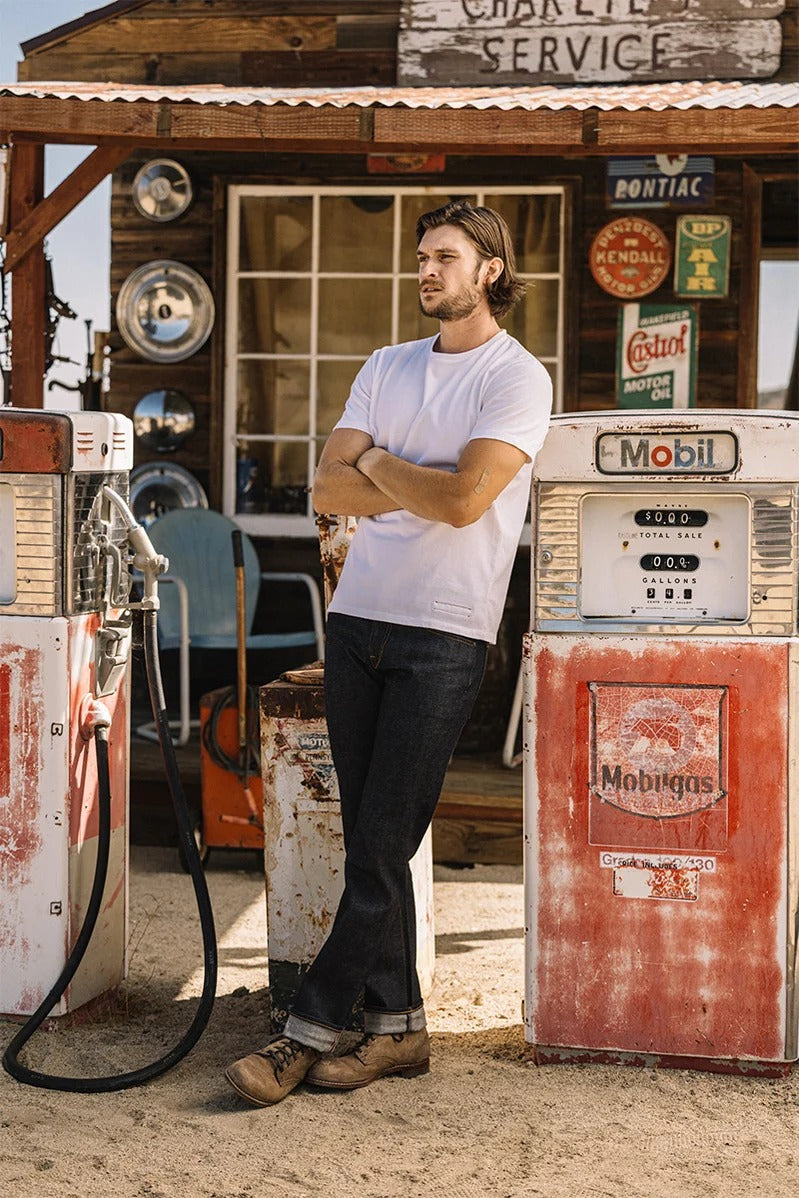 A man wearing a white T-shirt and Hiroshi Kato - The Hammer Straight / Indigo Raw 14oz Selvedge jeans stands with arms crossed between two vintage gas pumps in front of an old service station adorned with signs and hubcaps.