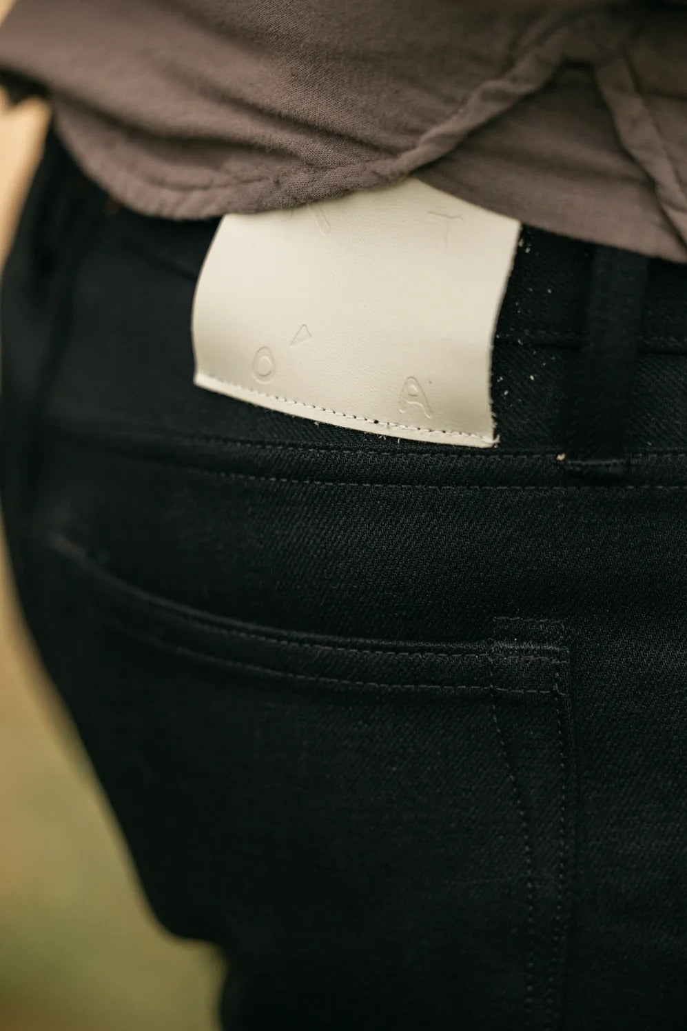 A close-up of a person wearing Hiroshi Kato - The Hammer Straight / Black Raw 14oz Selvedge jeans with a beige patch on the waistband and a grey shirt, highlighting the back pocket and part of the shirt hem—crafted from premium Japanese selvedge denim by Hiroshi Kato for timeless style and exceptional quality.