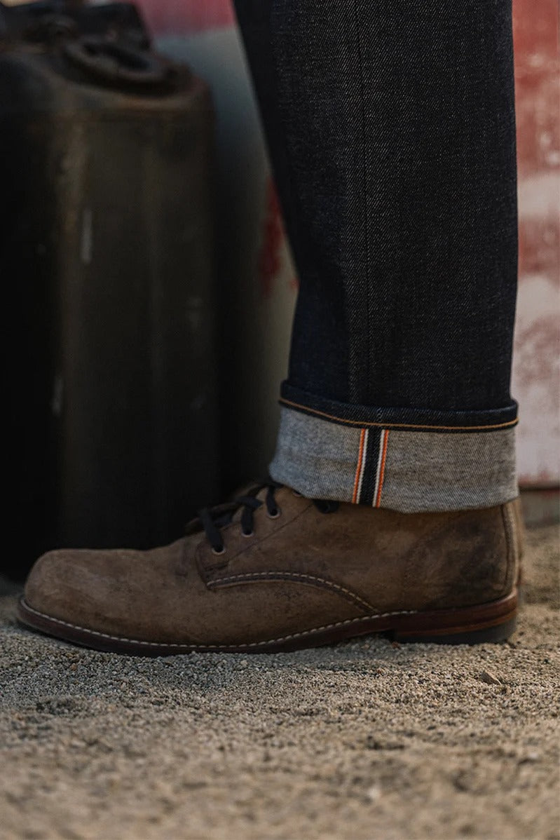 A close-up of a person’s lower leg wearing Hiroshi Kato - The Hammer Straight / Indigo Raw 14oz Selvedge jeans, featuring visible Japanese selvedge, paired with brown suede shoes and standing on sandy ground near a black container.