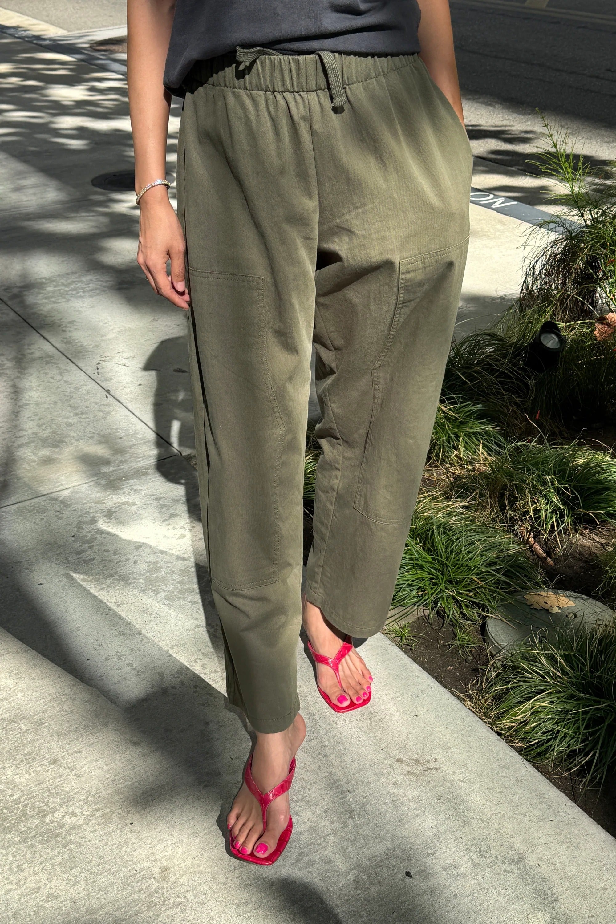 A person wearing Le Bon Shoppe - Gardener’s Pant in Army Green styled with a relaxed silhouette and a gray shirt stands on a sunlit sidewalk. They pair the look with bright pink sandals, while shadows and greenery line the edge of the sidewalk.