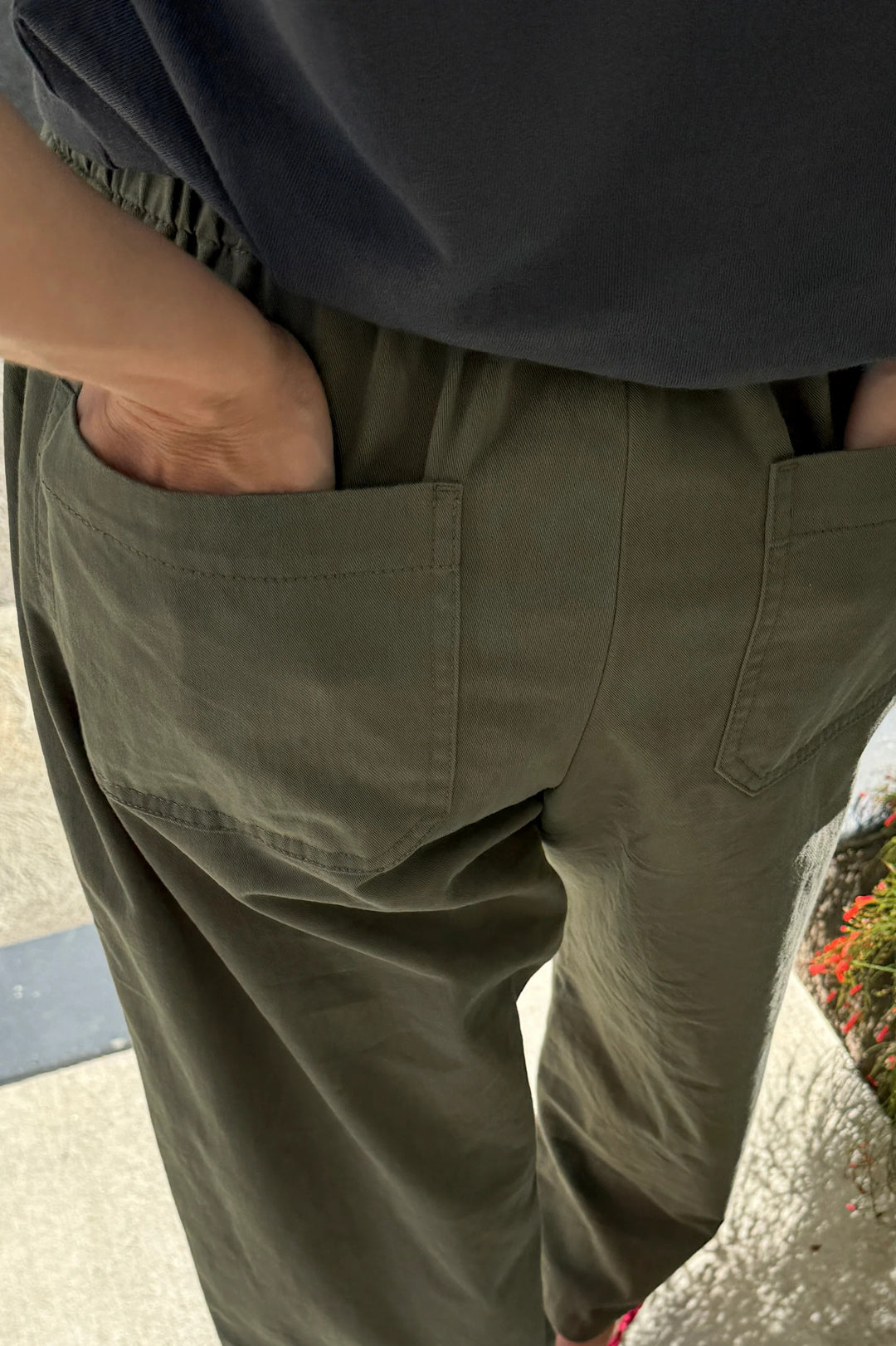 A person stands outdoors on a sidewalk next to flowers, wearing Le Bon Shoppe - Gardener’s Pant in Army Green. The relaxed silhouette of these pants, crafted from lightweight cotton-nylon fabric, adds comfort to this laid-back look. Only the lower torso and upper legs are shown, with both hands tucked into the back pockets.