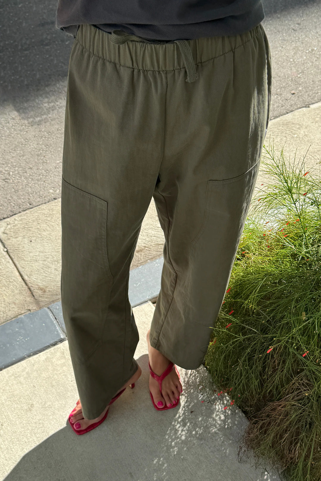 A person wearing Le Bon Shoppe - Gardener’s Pant in Army Green, designed with a relaxed silhouette from lightweight cotton-nylon, stands on a sidewalk near greenery with part of a road in the background. The image shows only the lower half of their body, paired with red sandals.
