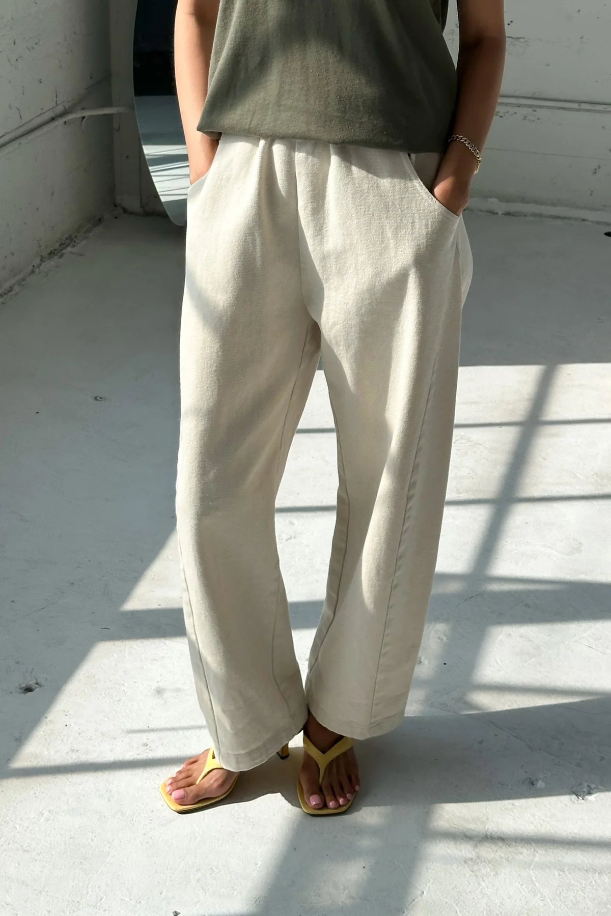 A person stands indoors in sunlight, wearing Le Bon Shoppe - Arc Pant / Natural—loose, light beige pants paired with a khaki green top and yellow sandals. Their hands are in their pockets; only their body from the shoulders down is visible.