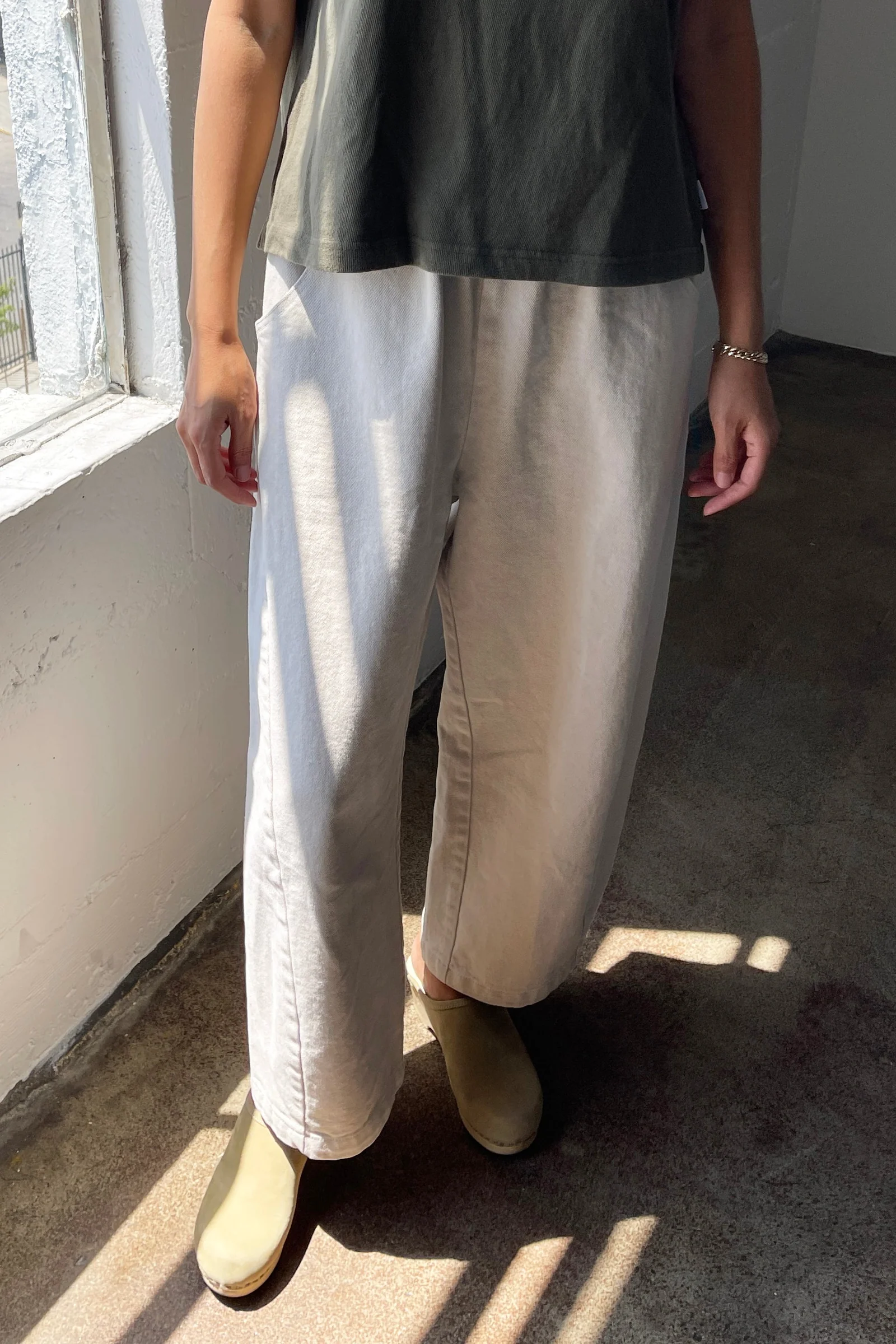 A person stands indoors by a sunlit window, wearing a dark short-sleeve shirt, Le Bon Shoppe - Arc Pant in Natural, olive green shoes, and a bracelet. Sunlight casts shadows on their clothes and the floor.