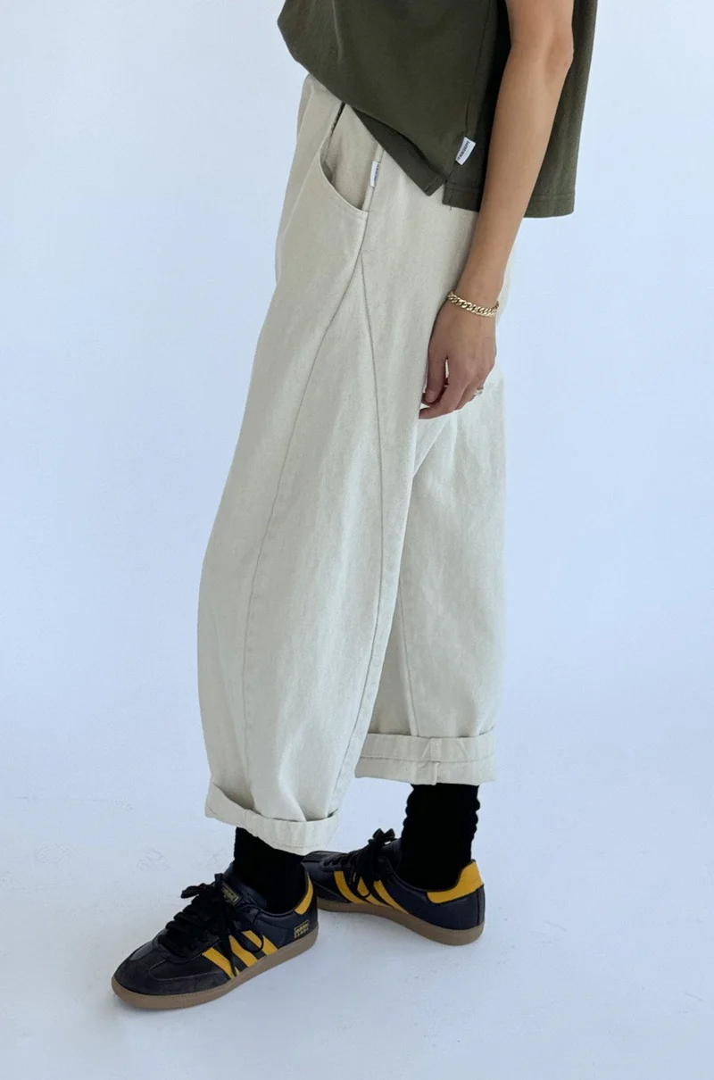 A person stands sideways against a plain light background wearing Le Bon Shoppe - Arc Pant in Natural, a dark green shirt, black socks, black and yellow sneakers, and a gold bracelet.