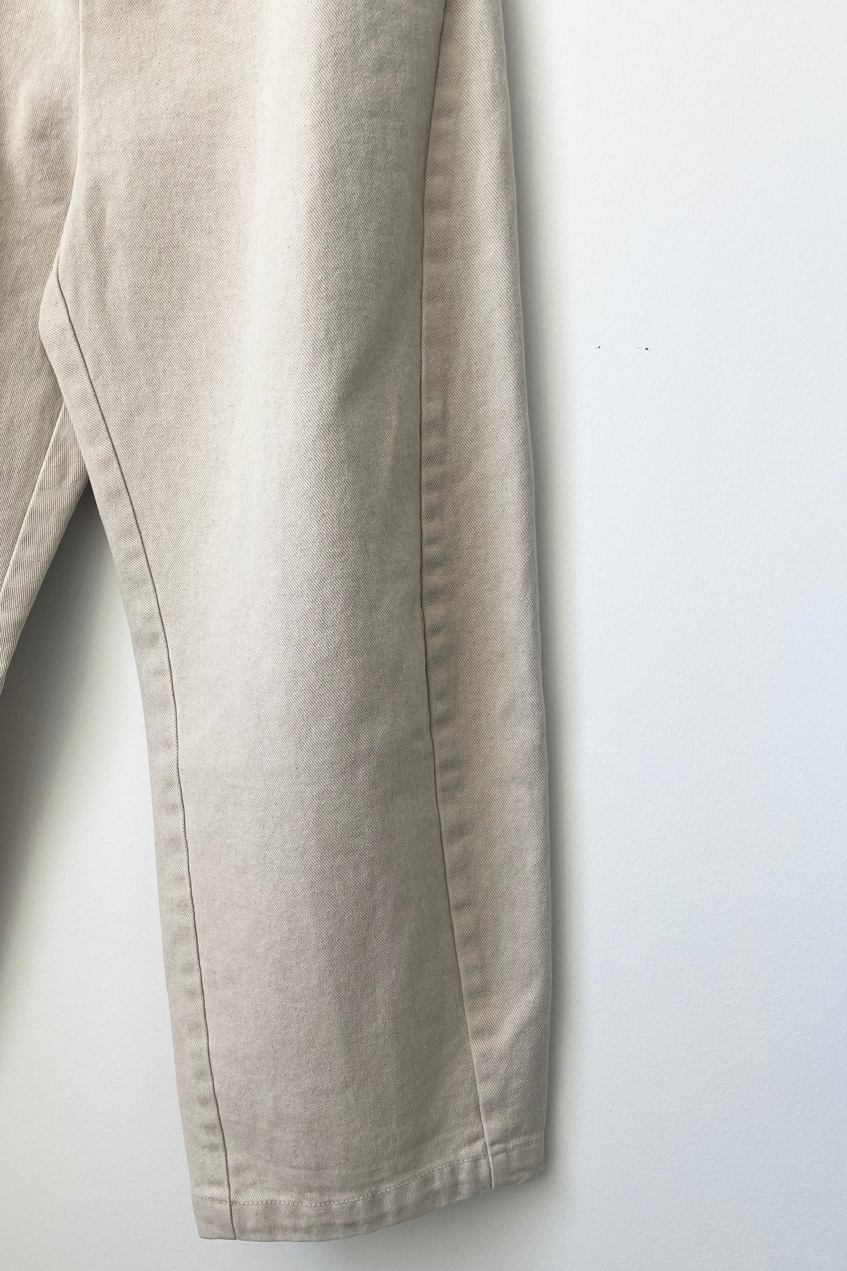 A close-up of the Le Bon Shoppe - Arc Pant in Natural, hanging against a plain white wall, showcases the detailed leg seam and the texture of its heavy-weight canvas.