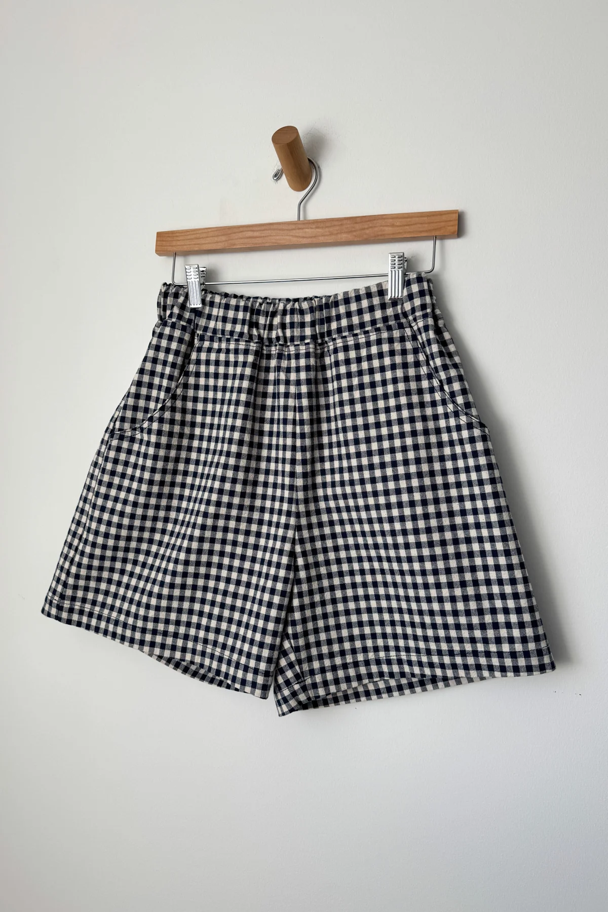 A wardrobe staple, the Le Bon Shoppe - City Short in navy gingham by Le Bon Shoppe is displayed on a wooden hanger against a plain white wall.