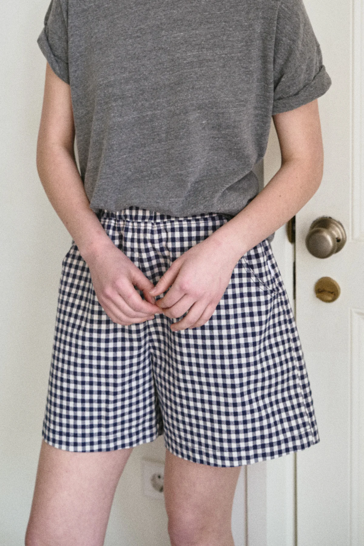 Wearing Le Bon Shoppe's City Short in Navy Gingham, this individual stands indoors with hands loosely clasped at the waist. Only their torso and legs are visible in this classic look from Le Bon Shoppe.