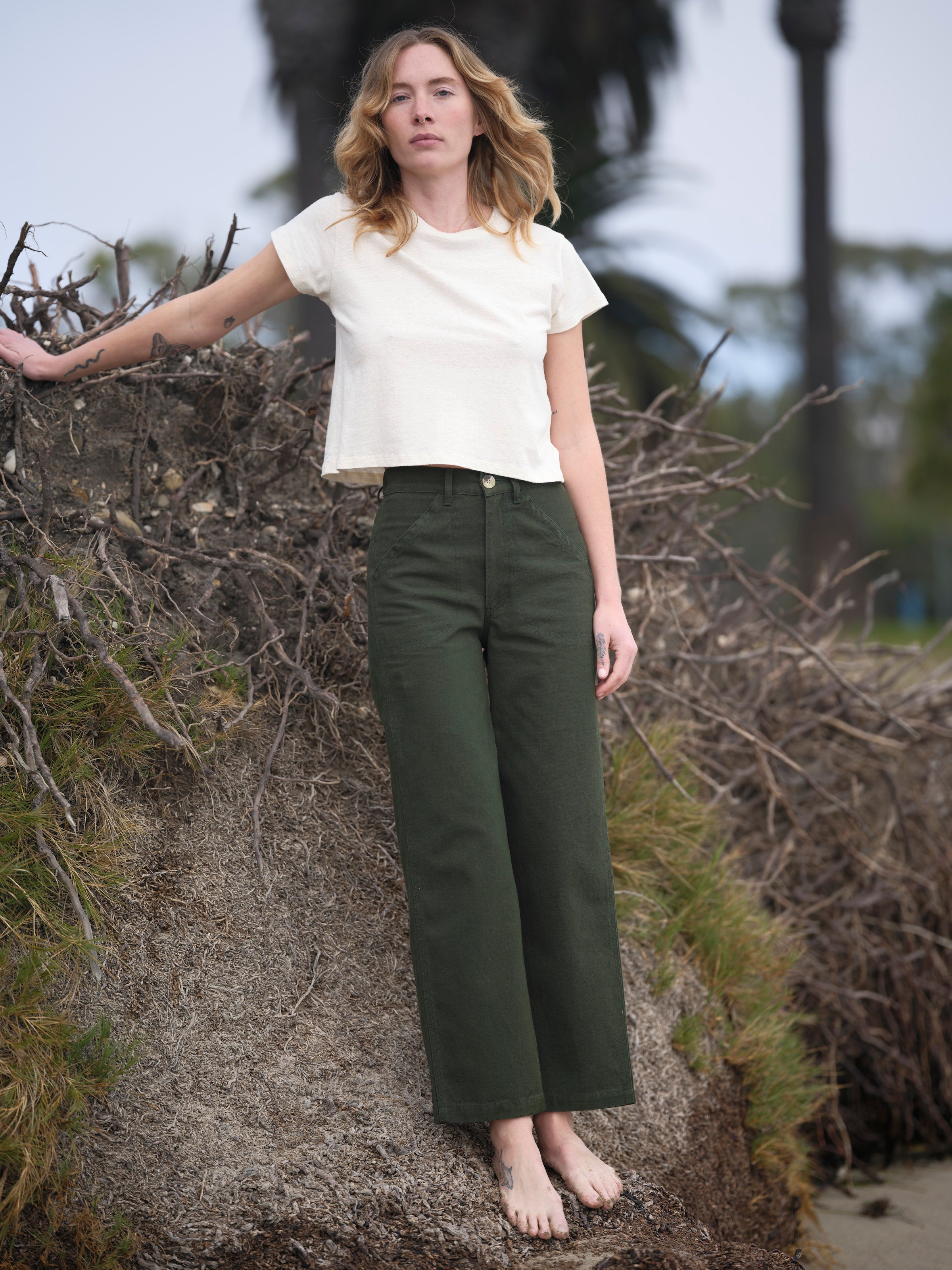 A woman with wavy blonde hair stands barefoot on sand near driftwood, wearing a white cropped t-shirt and Mollusk Painter Pant in Dark Rover Green by Mollusk. The background includes lush greenery and blurred palm trees.