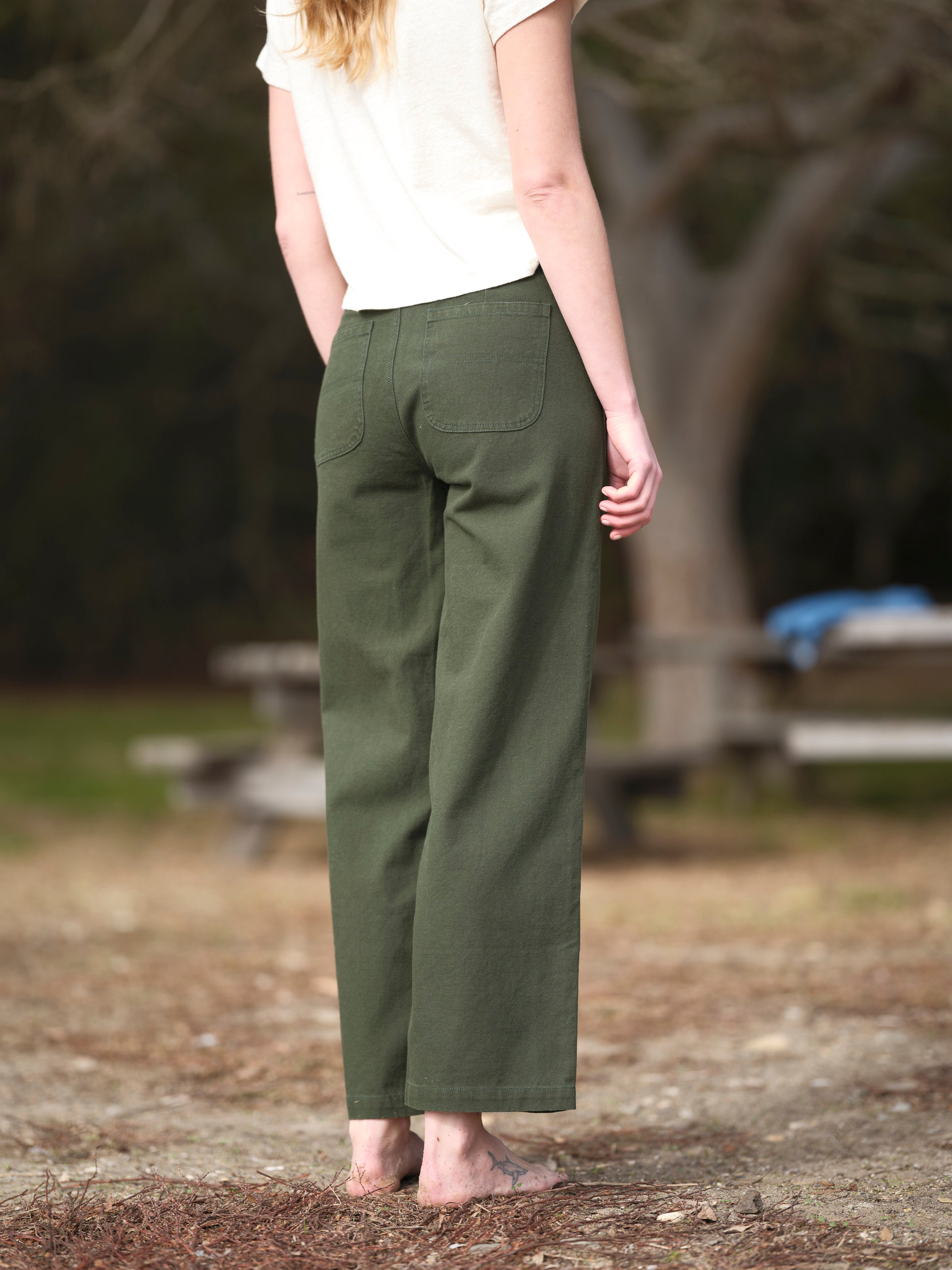 A barefoot person wearing a white short-sleeve top and Mollusk - Painter Pant / Dark Rover Green stands outdoors with their back to the camera, highlighting the stylish women’s bottoms from Mollusk in a relaxed natural setting.
