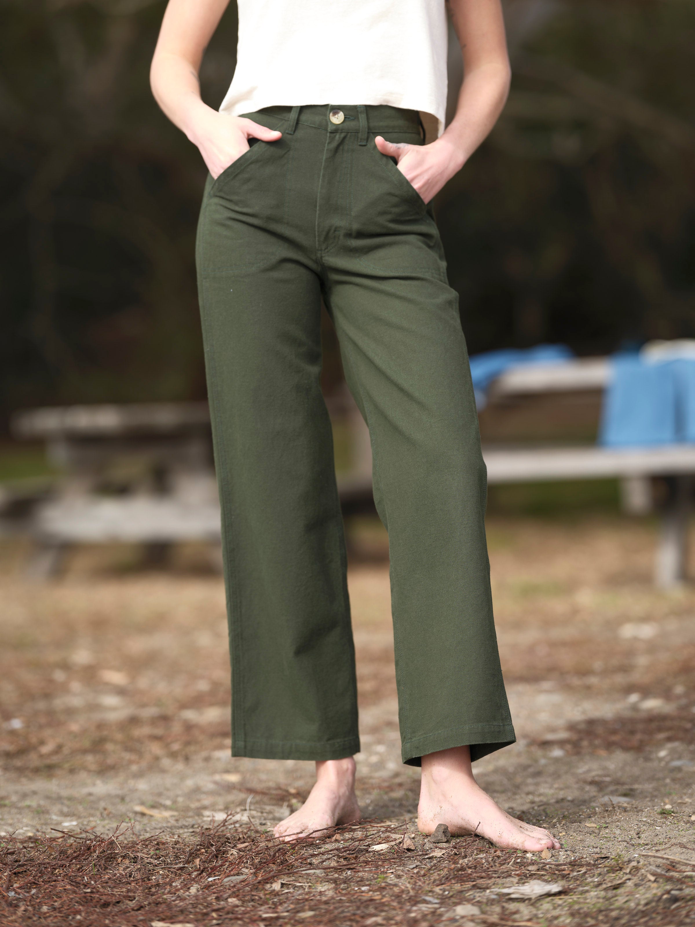 A person stands barefoot outdoors on dirt and grass, wearing Mollusk Painter Pant in Dark Rover Green and a white sleeveless top, hands in pockets. The background is blurred with a picnic table visible.