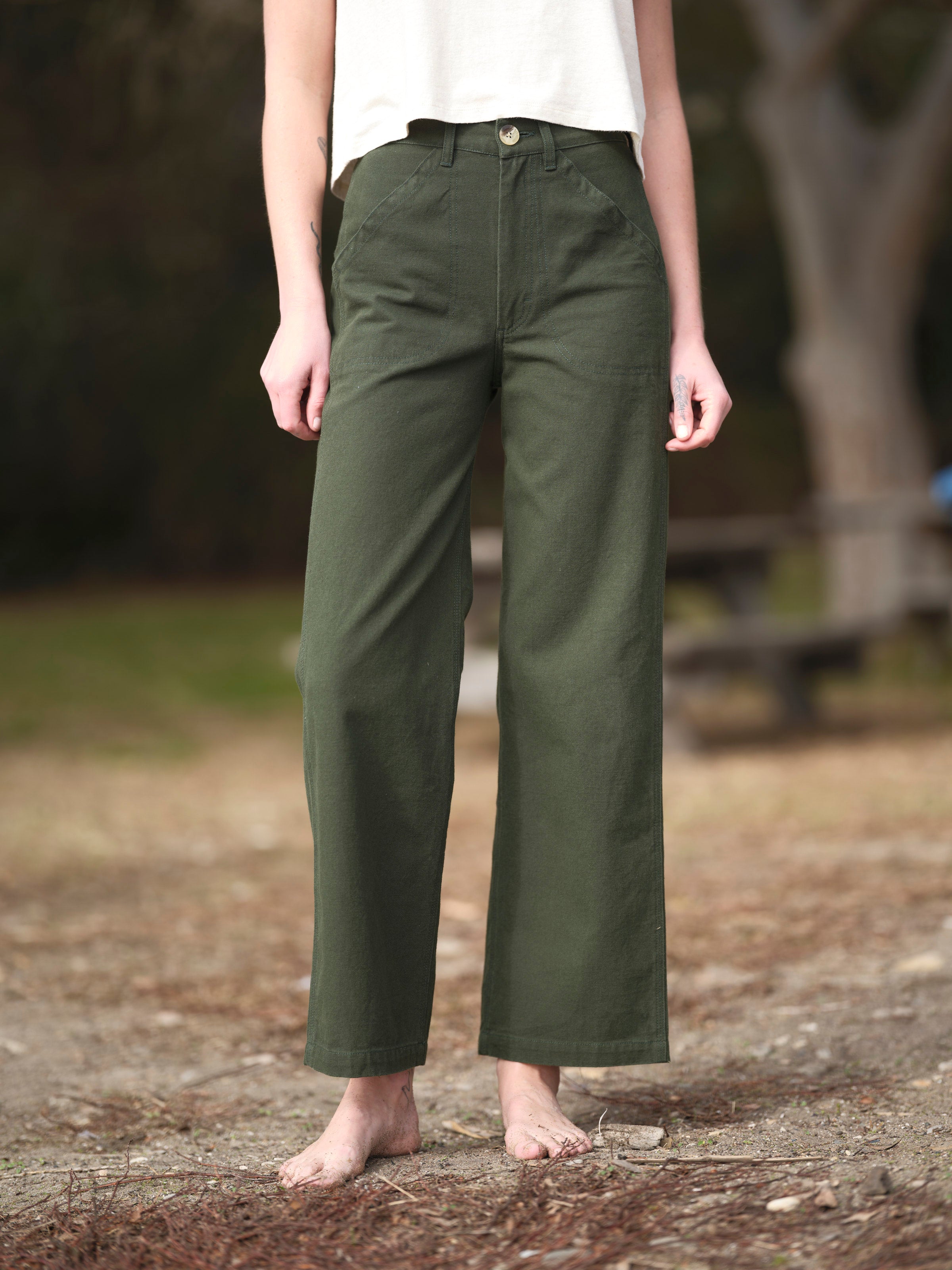 A person stands barefoot outdoors on dirt, wearing Mollusk - Painter Pant in Dark Rover Green and a white sleeveless top. The background is blurred with trees and a picnic table; the person's face is not visible.