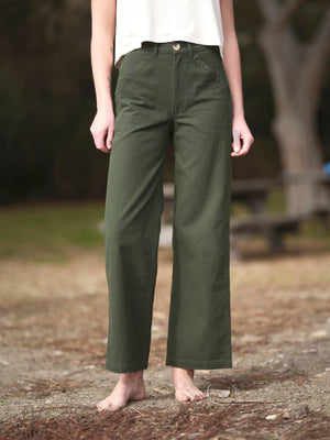 A person stands barefoot outdoors on dirt, wearing Mollusk - Painter Pant in Dark Rover Green and a white sleeveless top. The background is blurred with trees and a picnic table; the person's face is not visible.