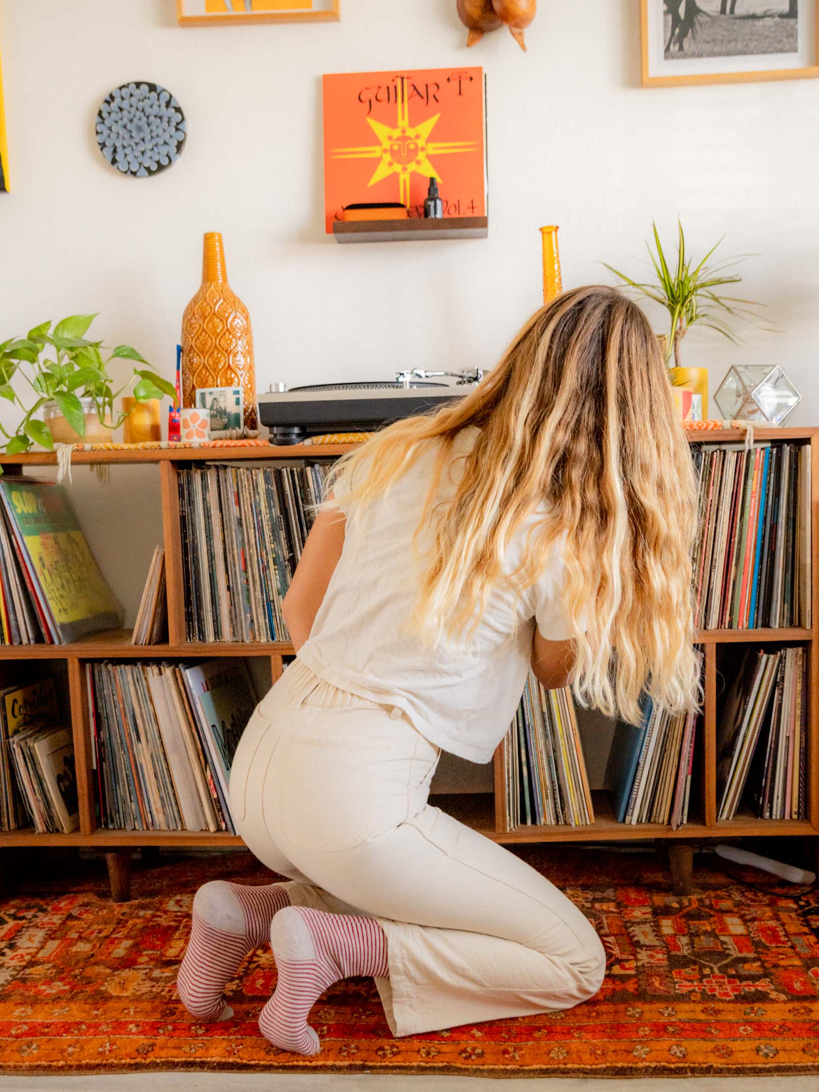 A woman with long blonde hair, wearing a white shirt and Mollusk - Painter Pant / Natural by Mollusk, kneels on a patterned rug while browsing a wooden record shelf in a cozy room filled with vinyl albums and plants.