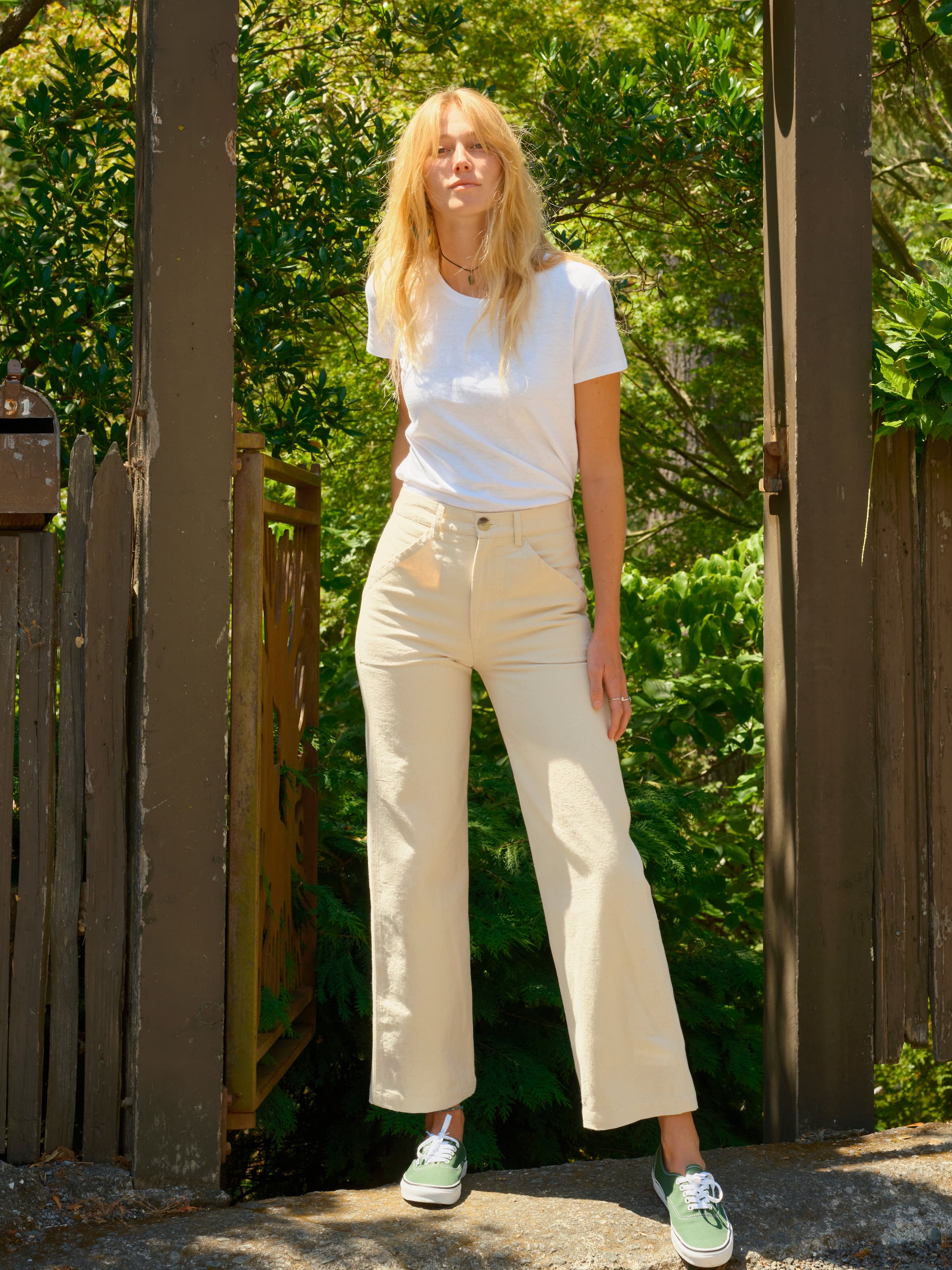 A woman with long blonde hair stands outside in sunlight by a wooden fence and greenery, wearing a white T-shirt, green sneakers, and Mollusk Painter Pant / Natural by Mollusk.