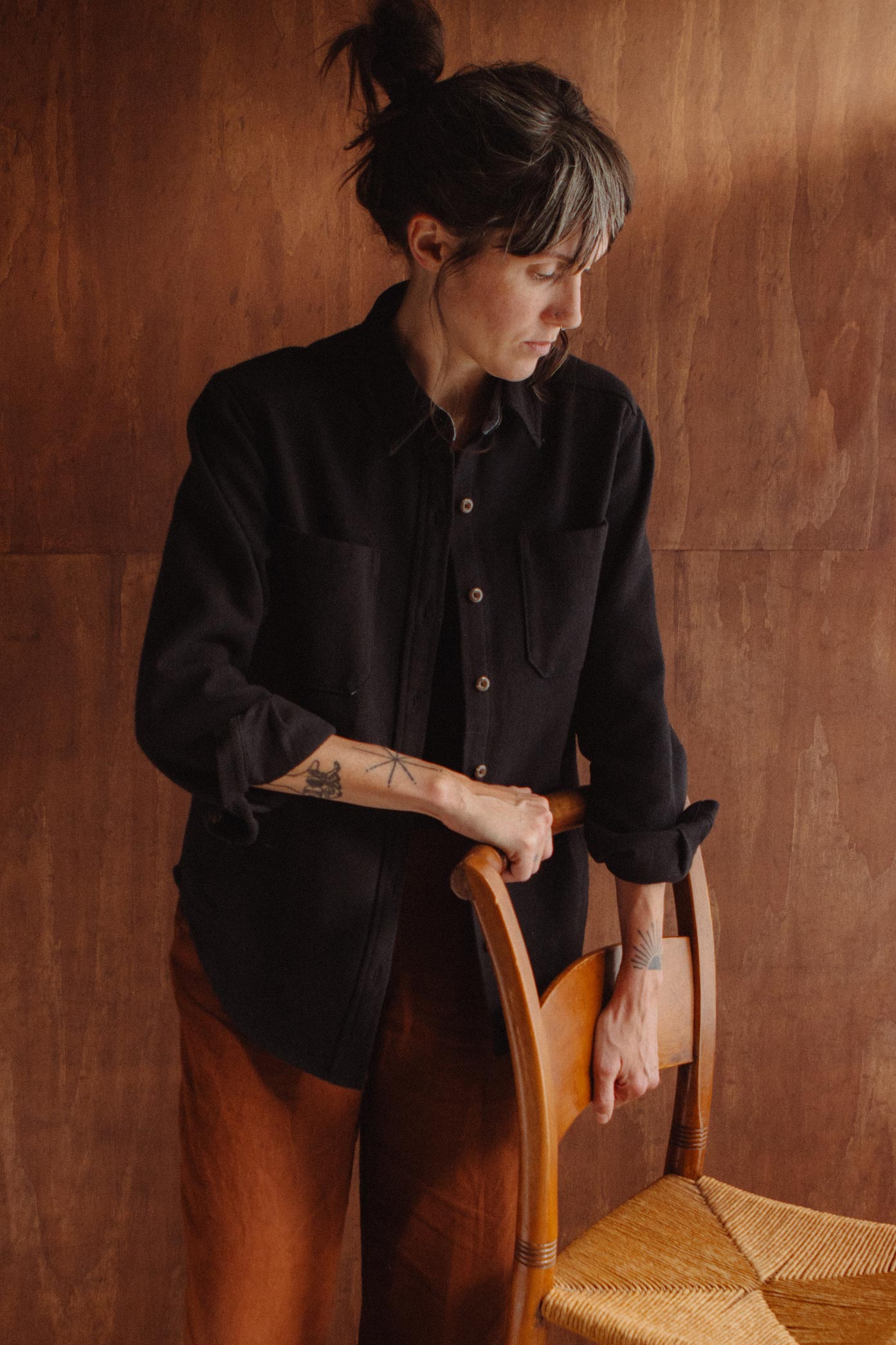A person wearing a dark recycled cotton Dawson Overshirt / Ink Stone stands with their hand resting on the back of a wooden chair.