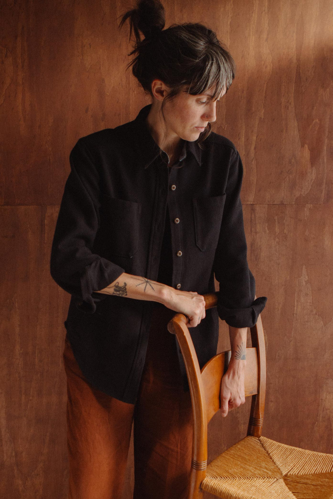A person wearing a dark recycled cotton Dawson Overshirt / Ink Stone stands with their hand resting on the back of a wooden chair.