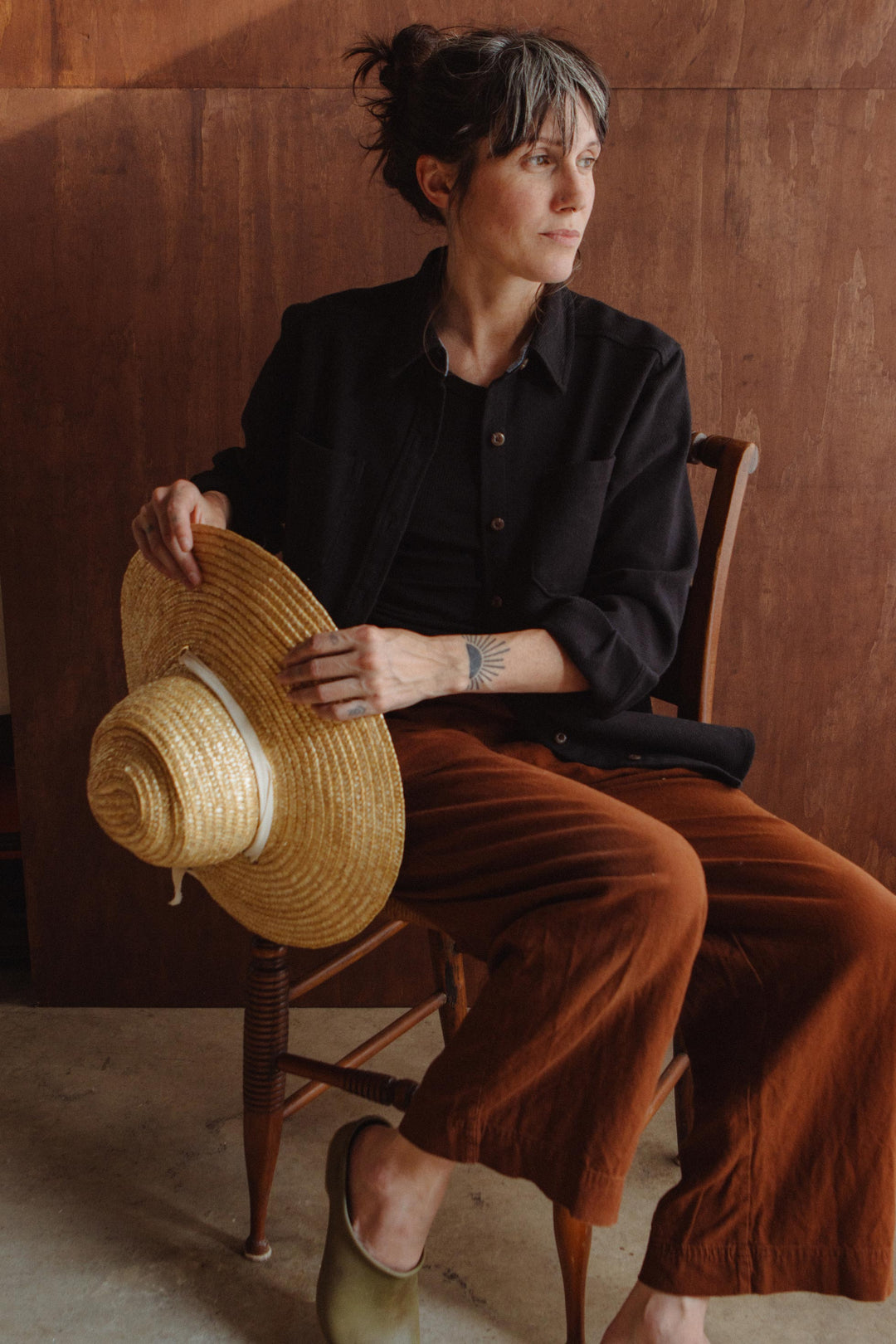 A person wearing a sustainable black recycled cotton Dawson Overshirt / Ink Stone and rust-colored trousers sits on a wooden chair, holding a straw hat.