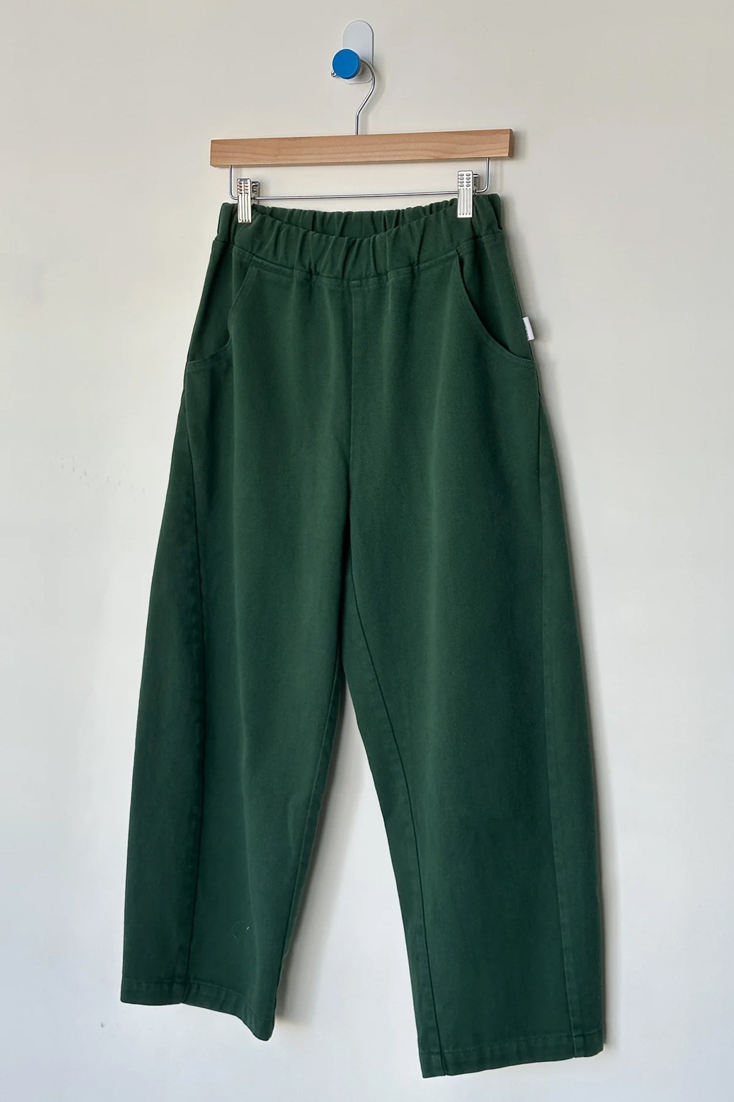 Le Bon Shoppe - Arc Pant / Pine: Dark green, high-waisted wide-leg denim pants with elastic waistband and side pockets, crafted from heavyweight denim, shown on a wooden hanger against a white wall by Le Bon Shoppe.