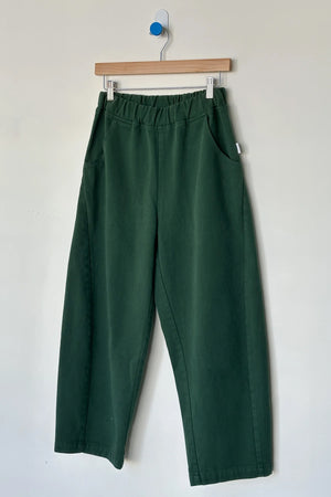 Le Bon Shoppe - Arc Pant / Pine: Dark green, high-waisted wide-leg denim pants with elastic waistband and side pockets, crafted from heavyweight denim, shown on a wooden hanger against a white wall by Le Bon Shoppe.