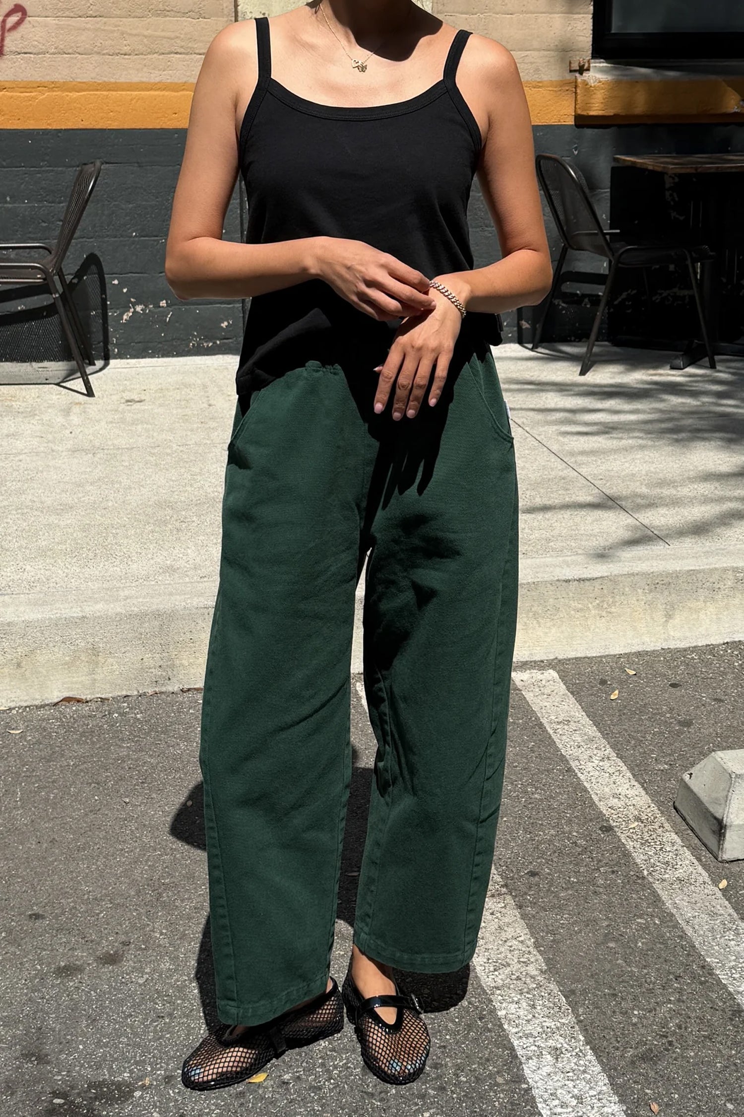 A person stands outdoors in sunlight, wearing Le Bon Shoppe - Arc Pant in Pine and a black tank top. Their hands are clasped, face partially out of frame, with shadows and outdoor chairs in the background.