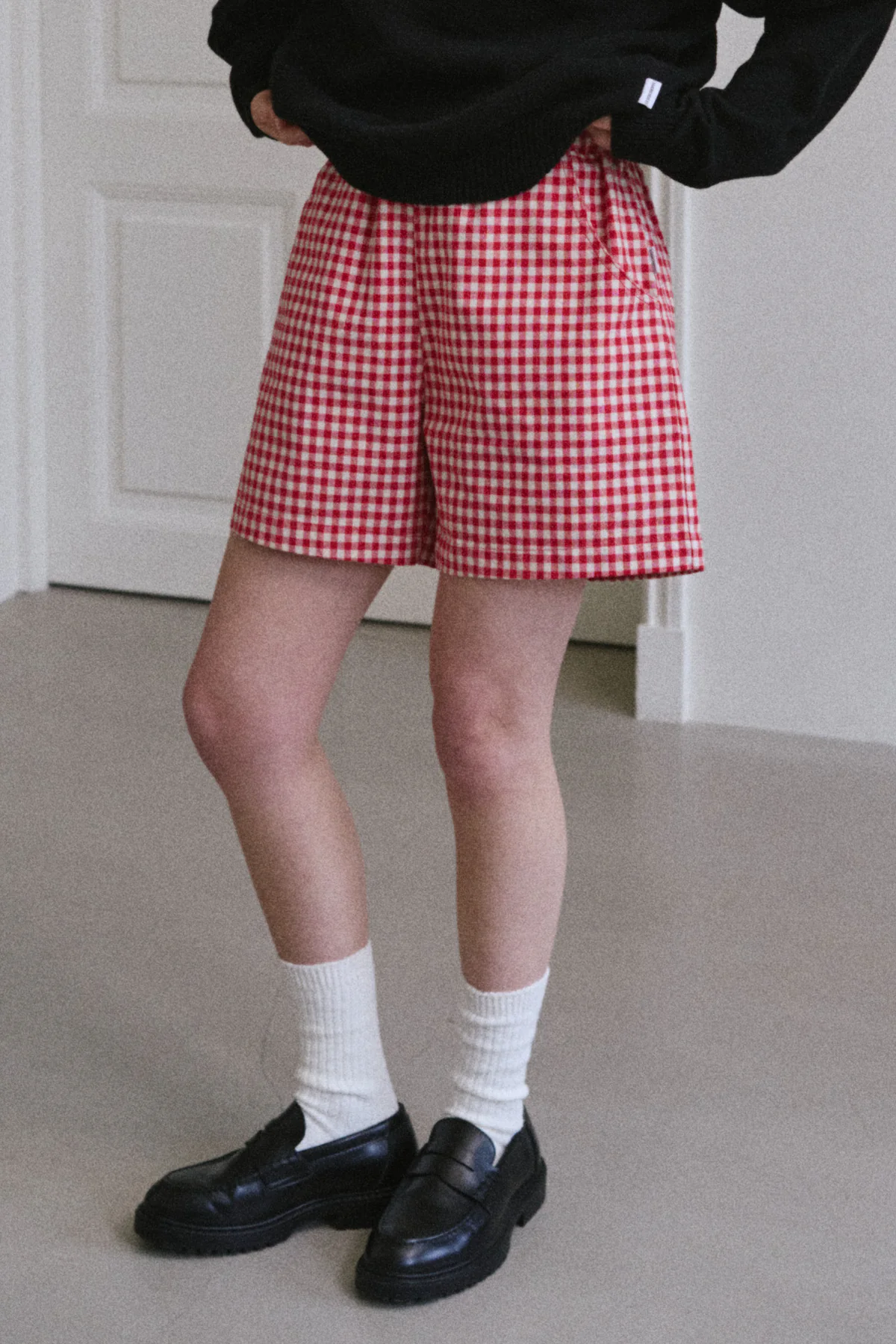 A person stands indoors wearing the Le Bon Shoppe - City Short in Red Gingham by Le Bon Shoppe, paired with a black sweater, white socks, and black loafers. Their hands are in their pockets, and only their lower body is visible.