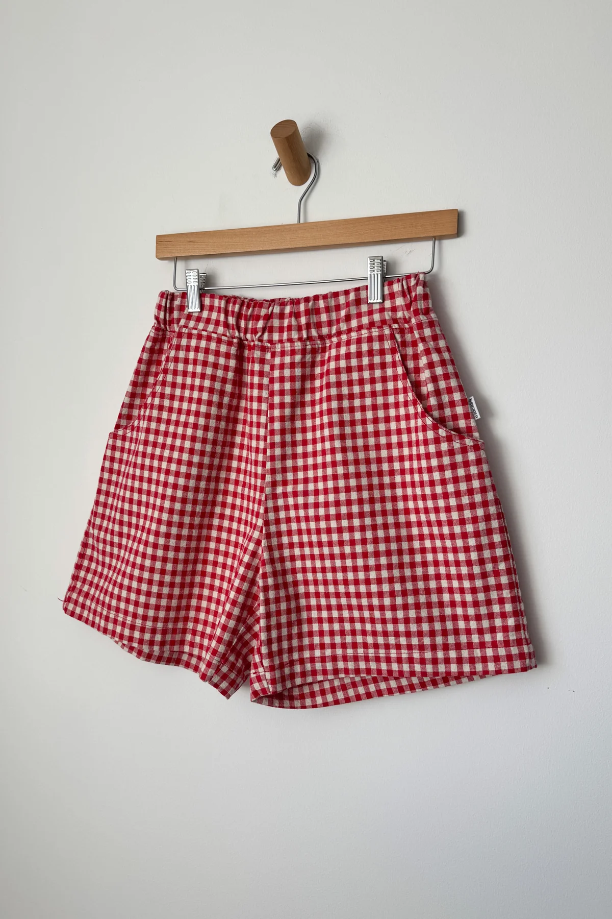 Le Bon Shoppe - City Short / Red Gingham: High-waisted shorts with side pockets and a relaxed fit, displayed on a wooden hanger against a plain white wall.