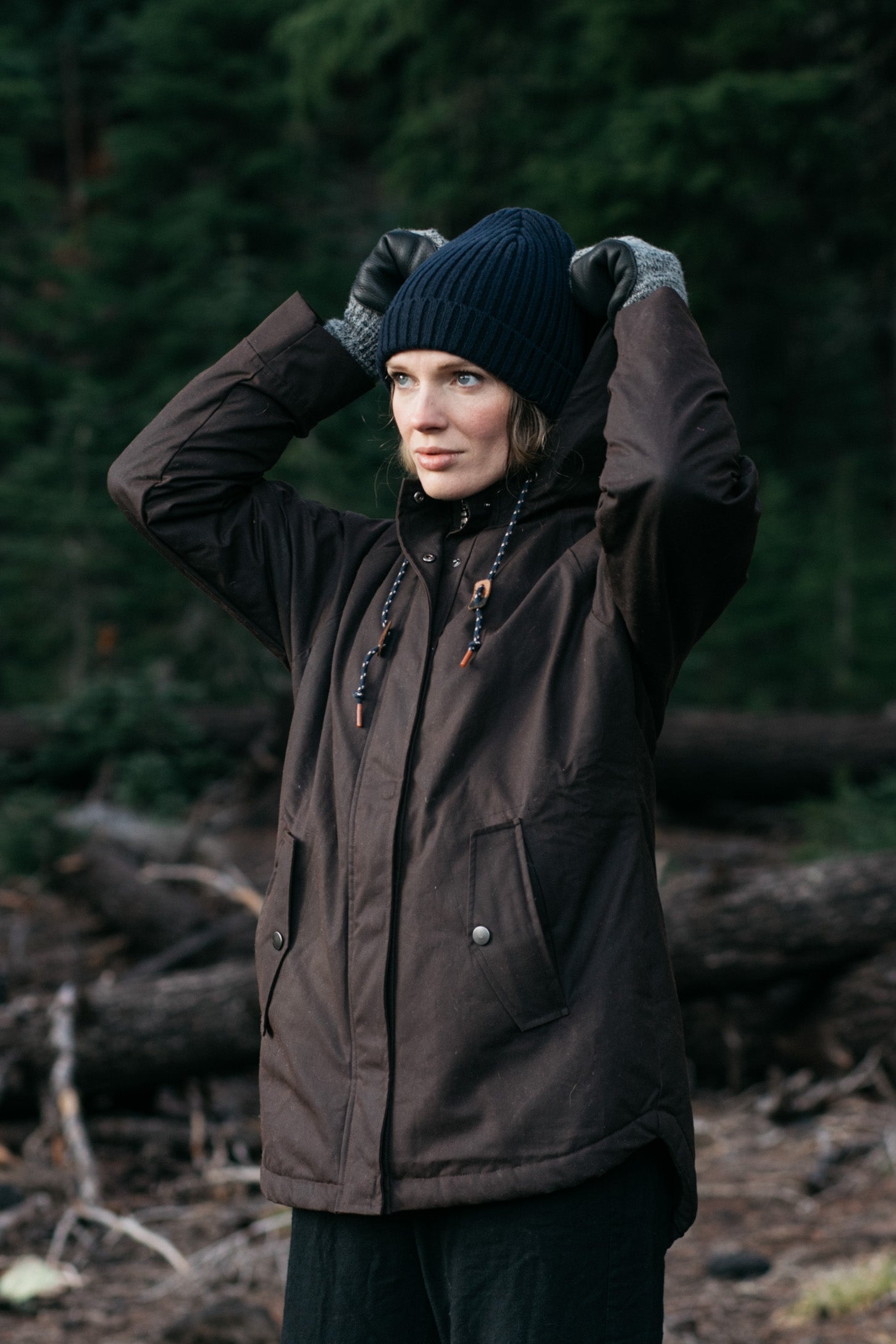 A person wearing the Bridge & Burn Sequoia Parka in Umber Waxed Canvas, black gloves, and a navy knit hat stands in a forest, adjusting their hat while looking into the distance.