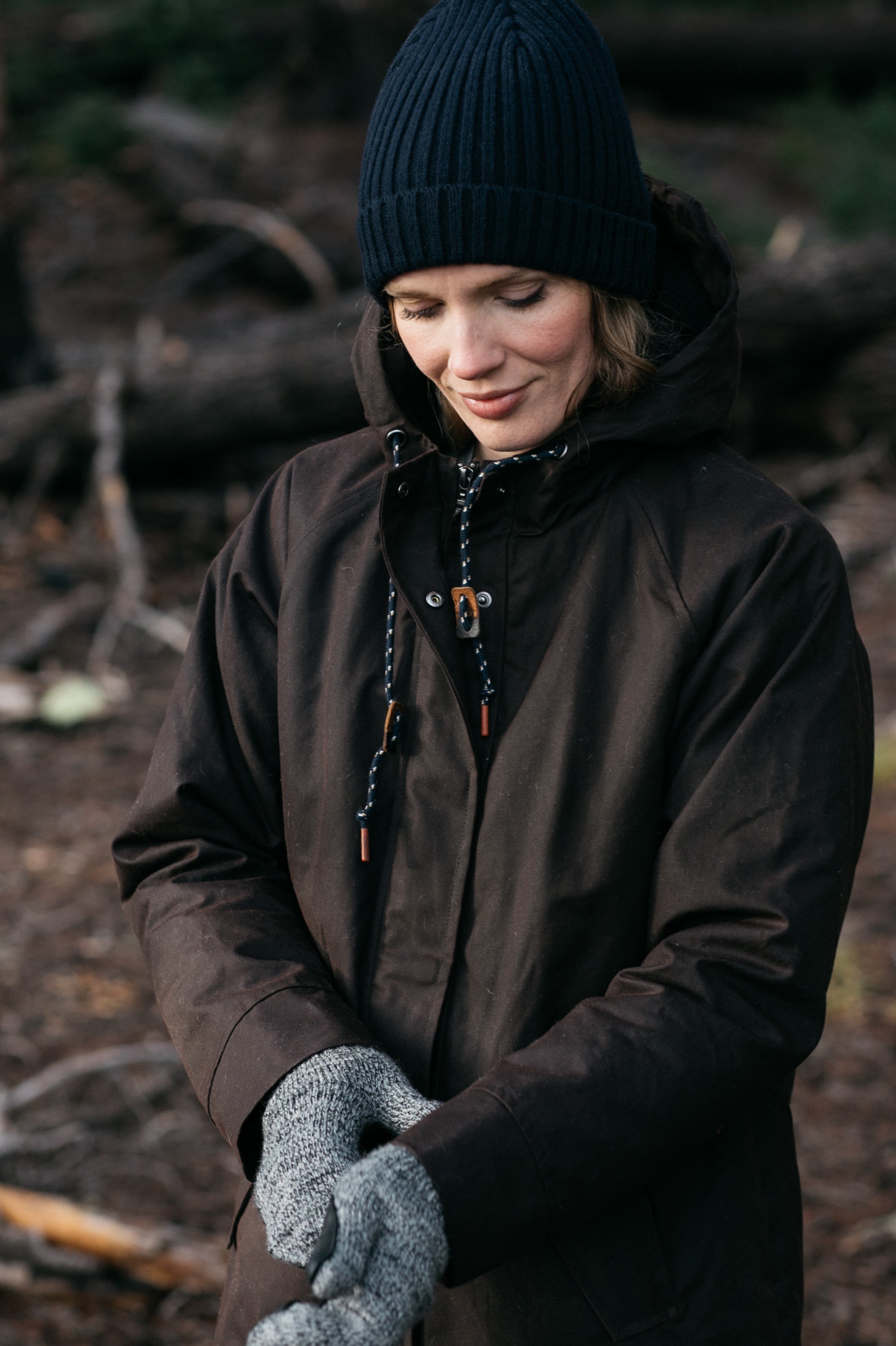 A person in a black beanie, gray knit gloves, and the Bridge & Burn Sequoia Parka in Umber Waxed Canvas stands outdoors among fallen branches and trees, looking down with a slight smile.
