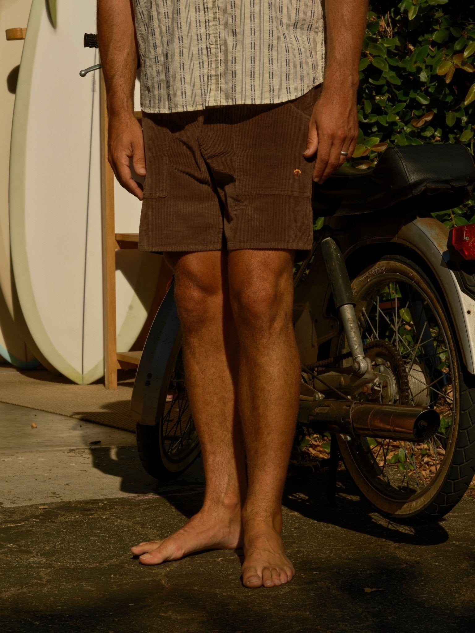 A barefoot man in Mollusk - Men's Shroom Short / Chocolate stands next to a bike and surfboard, with greenery and sunlight casting shadows in the background.