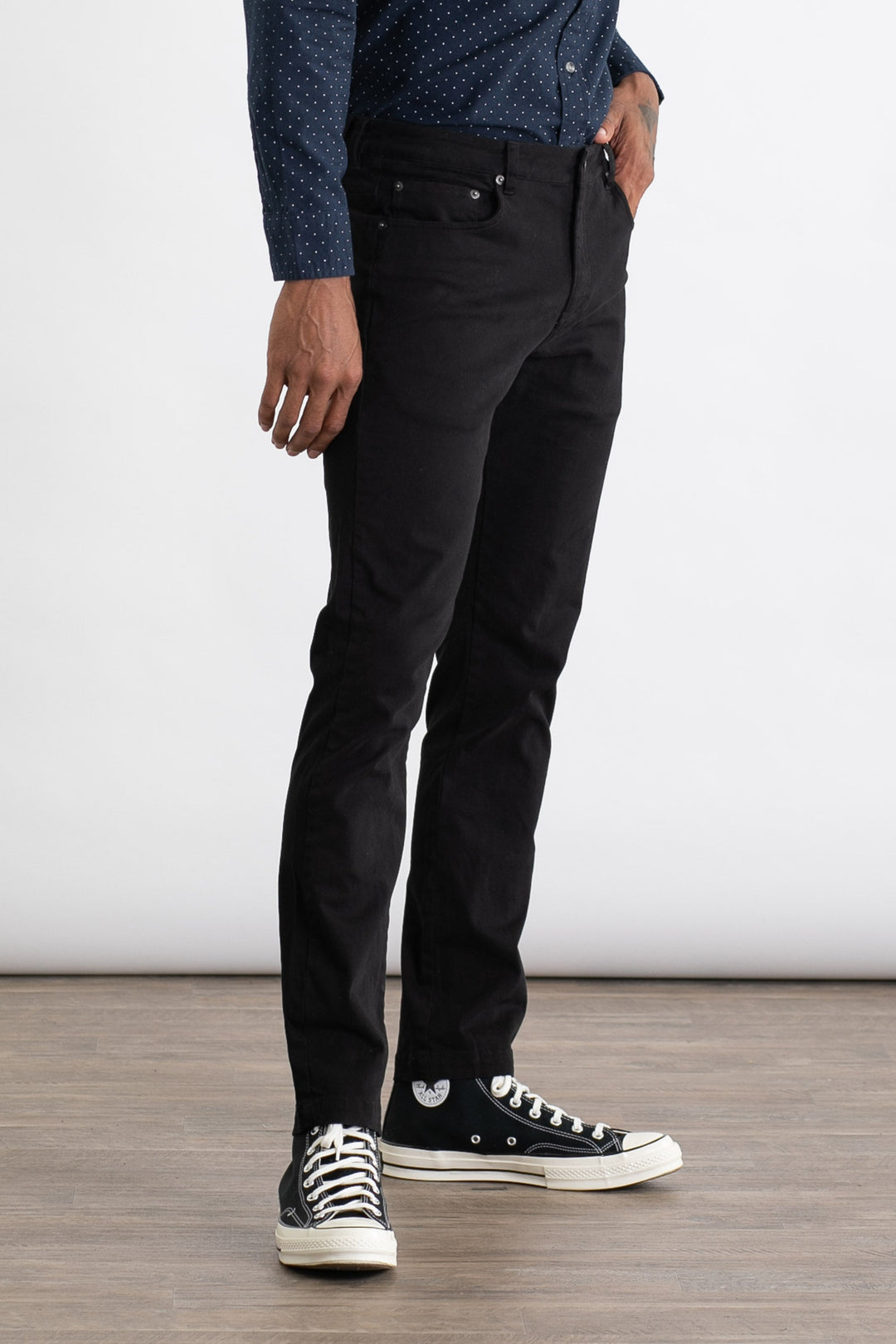 A person wearing a navy blue patterned shirt, Bridge & Burn Bradley Slim Pant in black, and black high-top Converse sneakers stands on a wooden floor against a plain white background.