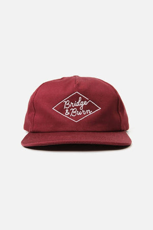 The Bridge & Burn Diamond Head Cap in Burgundy is a maroon baseball cap made from durable cotton twill, featuring white embroidered “Bridge & Burn” text inside a diamond-shaped outline on the front. It has a flat brim and a leather adjustable strap at the back for a comfortable fit.