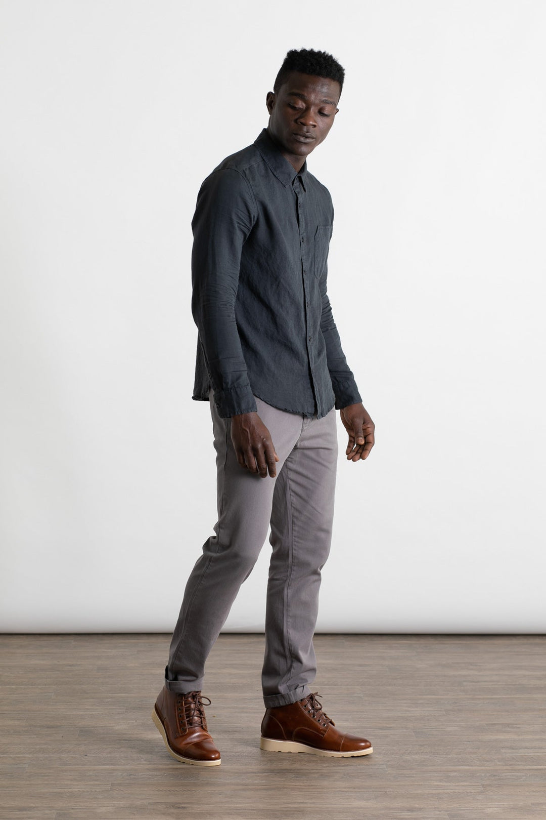 A man stands on a wooden floor, wearing Bridge & Burn Bradley Slim Pant in grey, styled with a dark button-up shirt and brown lace-up boots. He looks downward with his left hand at his side, set against a plain white background.