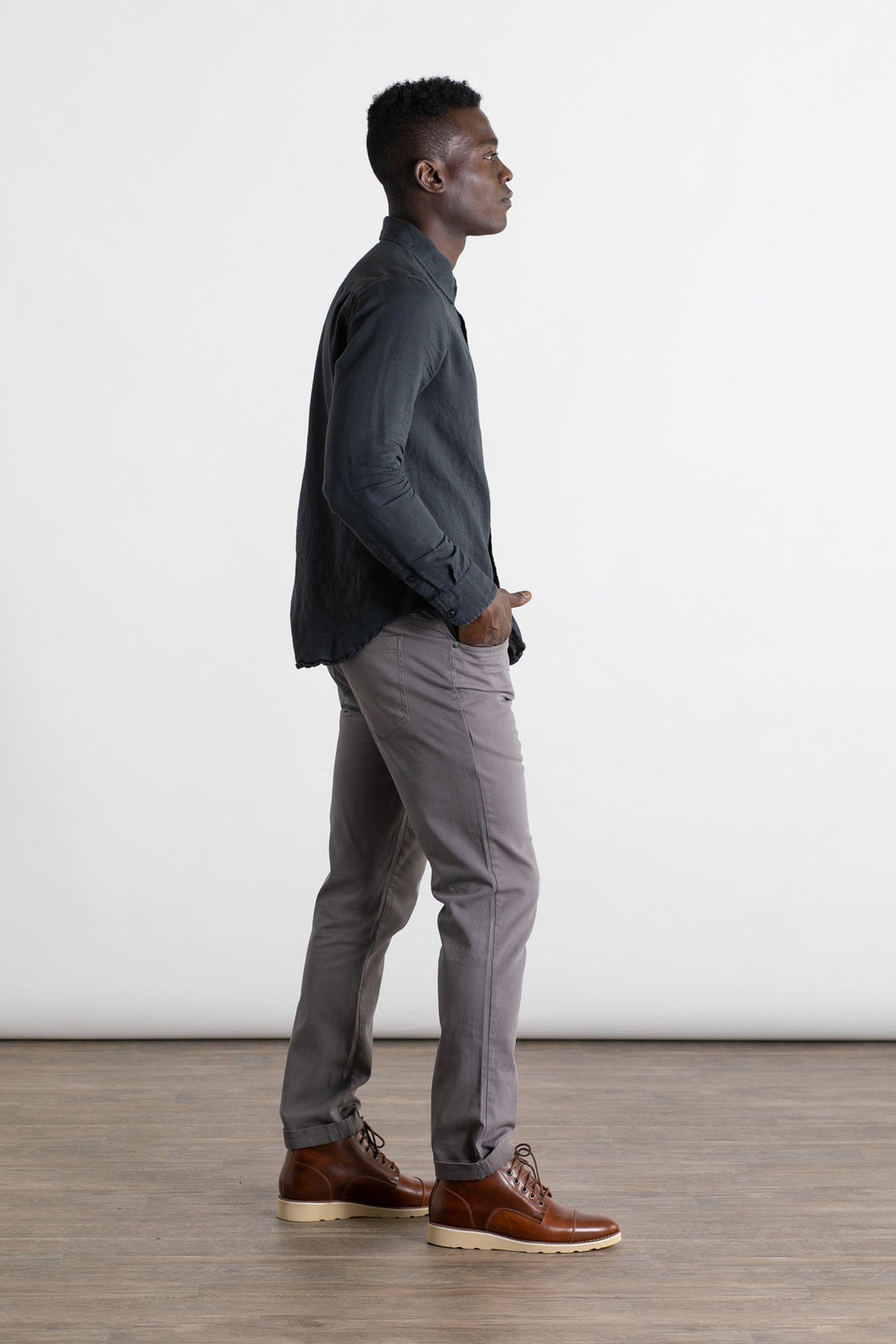 A man stands in profile against a white background, wearing a dark long-sleeve shirt and Bridge & Burn's Bradley Slim Pant in grey—a modern 5-pocket style—paired with brown leather shoes. One hand rests in his pocket as he looks ahead.