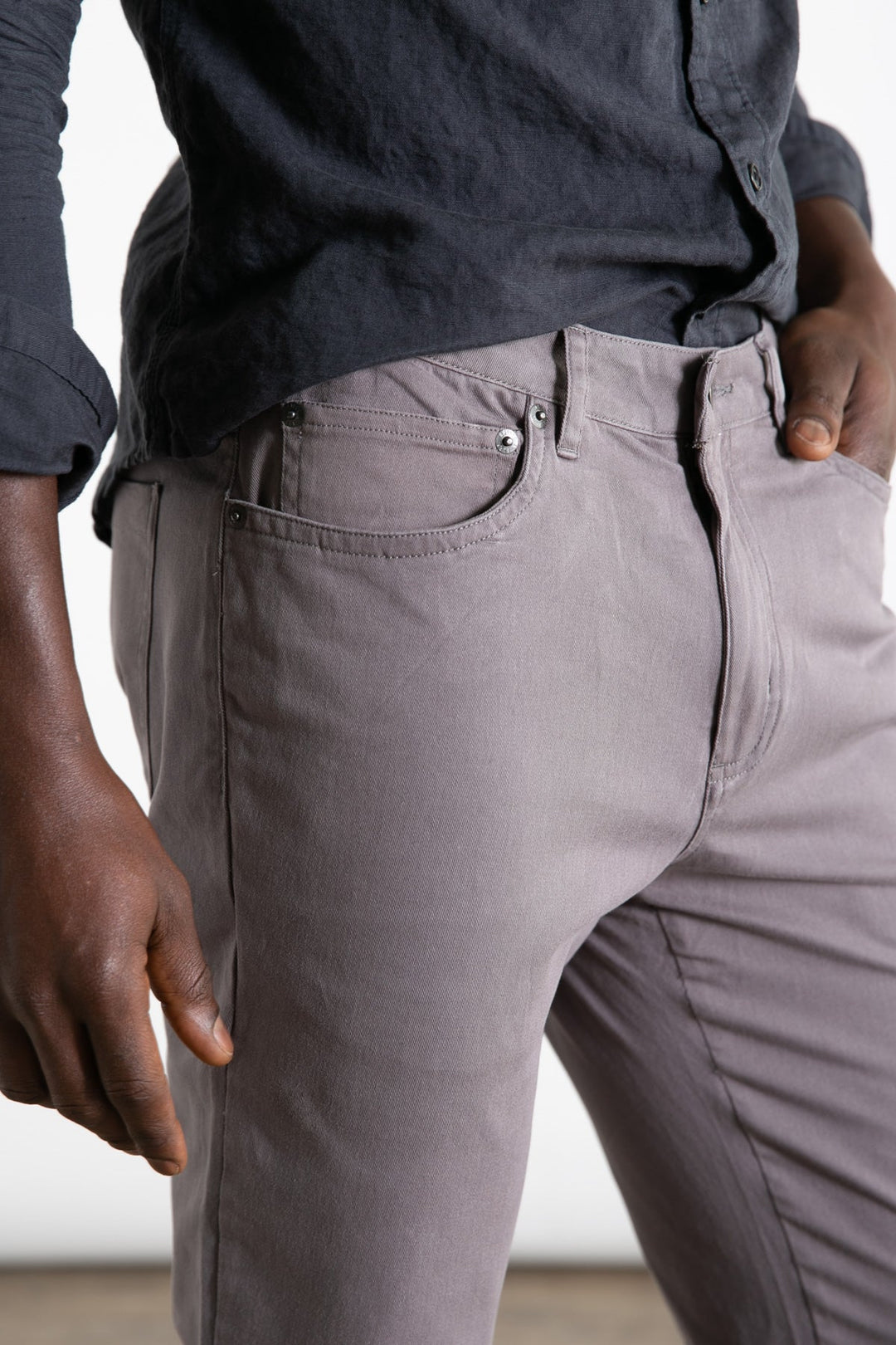 A person models the Bridge & Burn Bradley Slim Pant in grey, featuring a 5-pocket design. Styled with a dark long-sleeve shirt, they stand relaxed with one hand in their pocket; only torso and upper legs are visible.