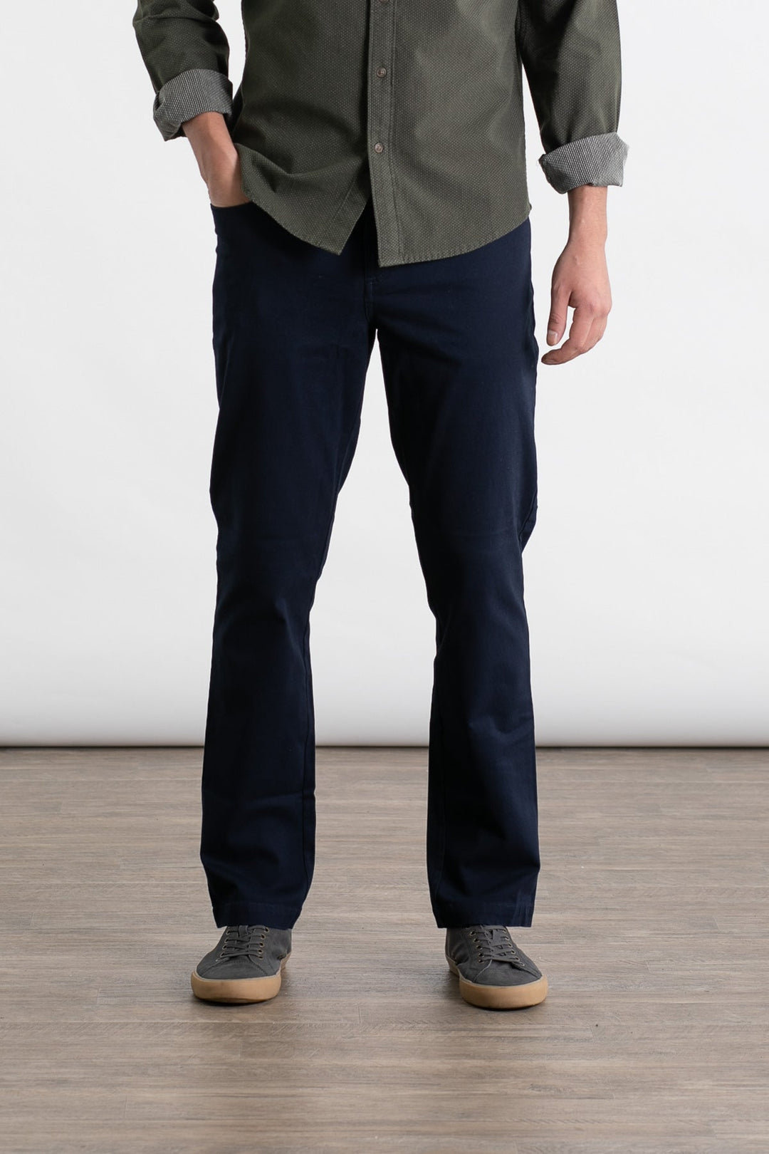 A person stands indoors on a wooden floor wearing Bridge & Burn’s Polk Pant in Navy, a green button-up shirt with rolled sleeves, and gray sneakers. The person's head is not visible in the image.