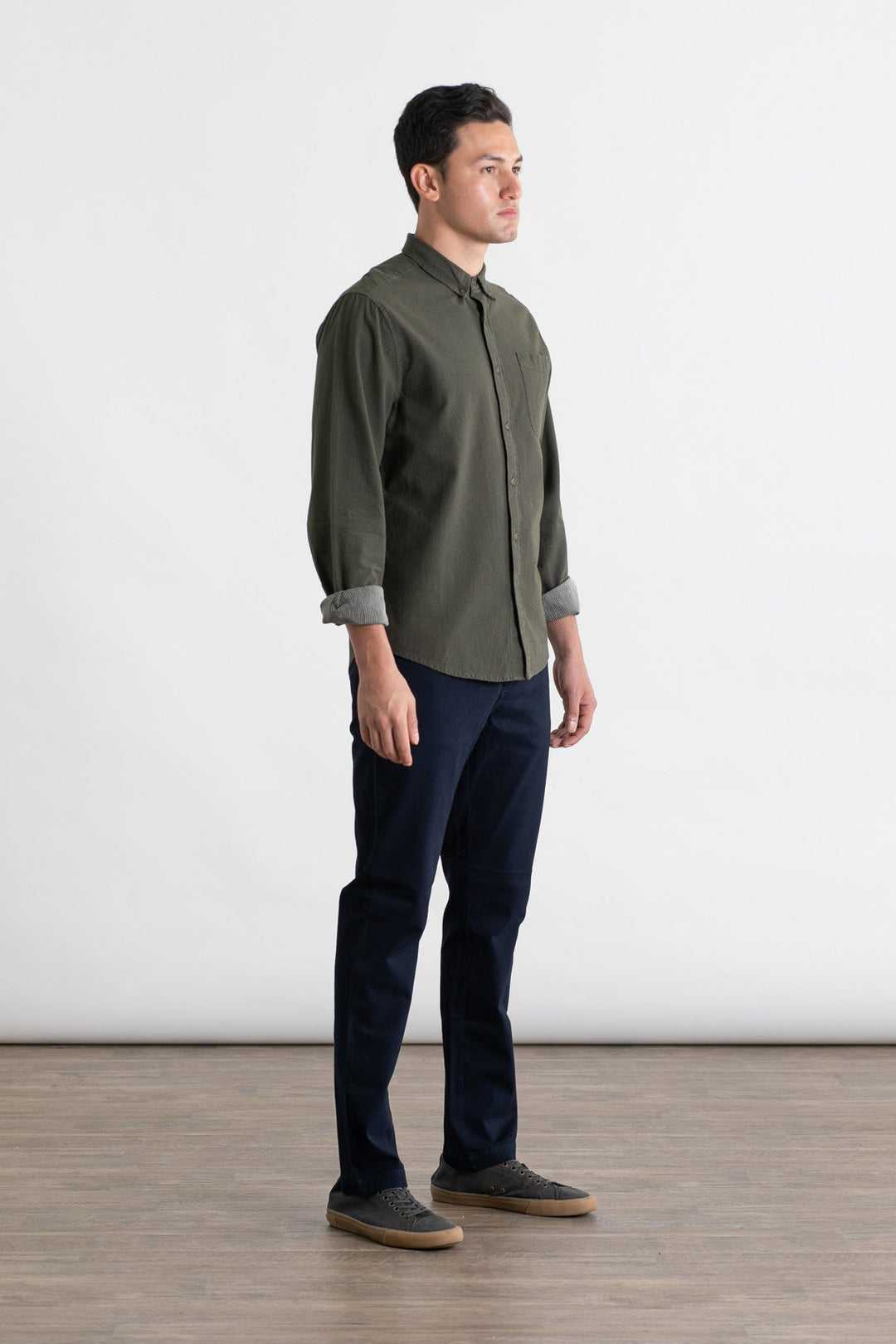 A man stands indoors on a wooden floor against a plain white background, wearing the Bridge & Burn Polk Pant in Navy, a dark green button-up shirt, and gray sneakers. He faces slightly right with a neutral expression.