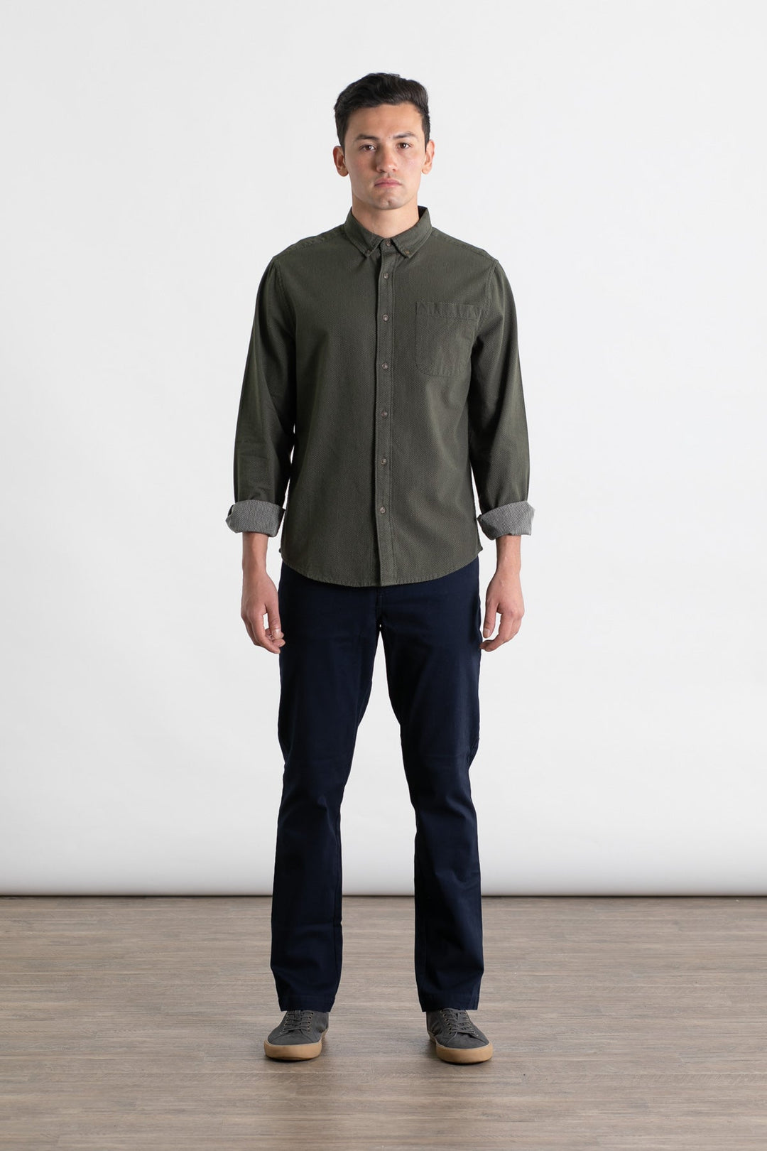 A man stands indoors on a wooden floor against a white background, wearing a dark green button-up shirt, gray sneakers, and Bridge & Burn’s Polk Pant in navy. His arms are relaxed at his sides as he faces forward.