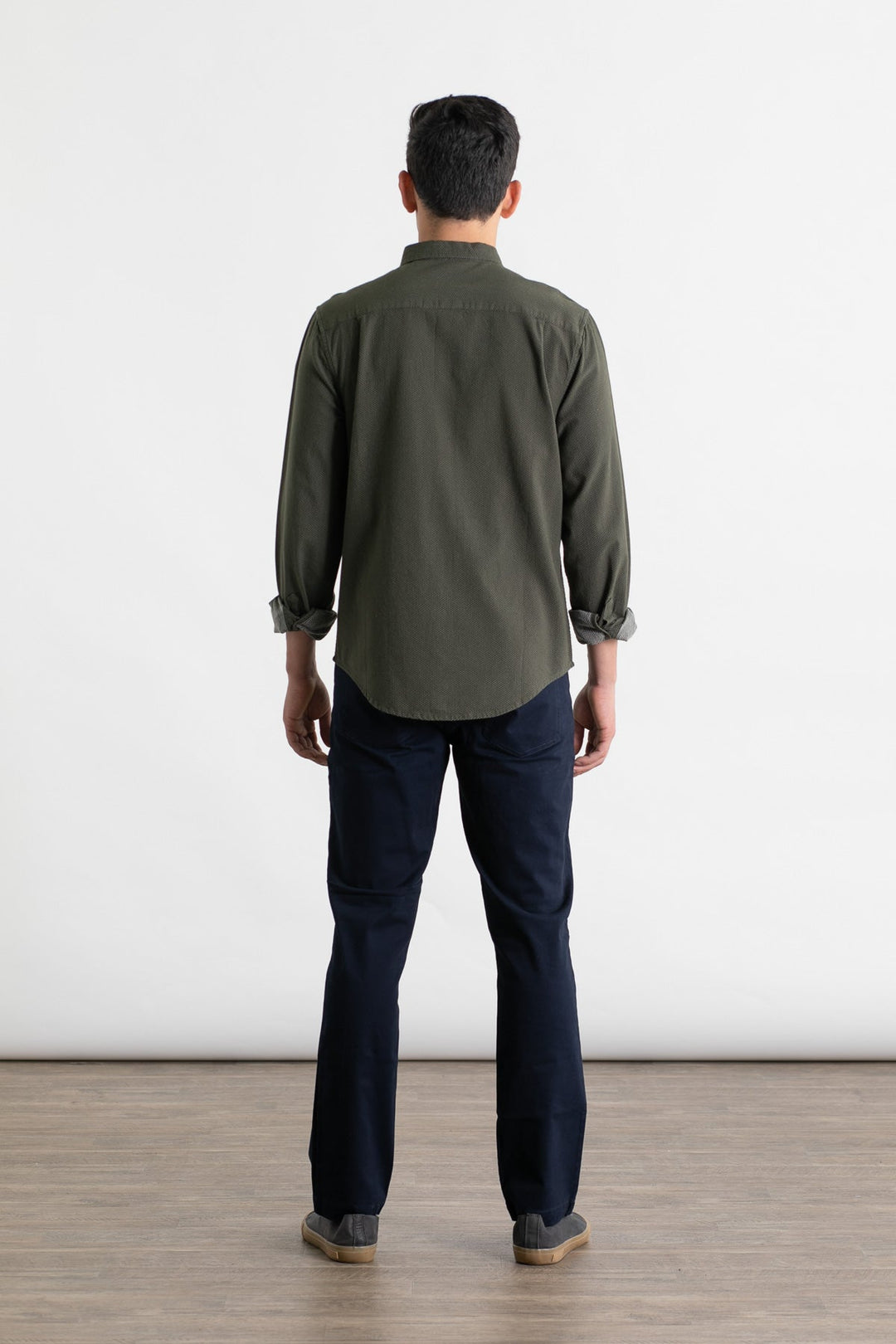 A man stands indoors with his back to the camera, wearing a long-sleeved olive green shirt and Bridge & Burn Polk Pant in Navy, paired with brown shoes, against a plain white background and light brown floor.