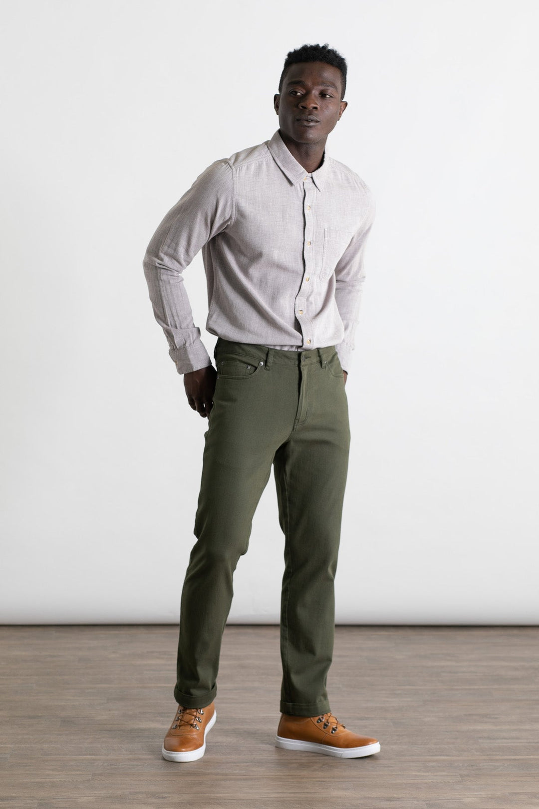 A man stands indoors on a wood floor, wearing a light gray button-up shirt, tan sneakers, and Bridge & Burn's Polk Pant in olive. He faces forward with hands in his pockets against a plain white background.