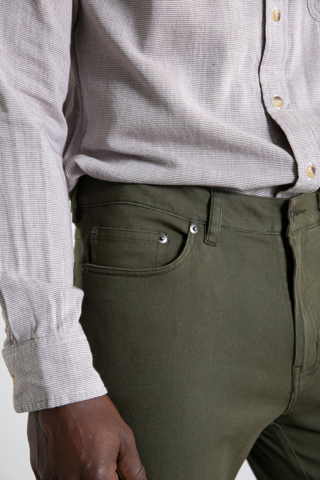 A close-up of someone wearing the Bridge & Burn Polk Pant in olive, paired with a light button-up shirt, showing details of the shirt cuff, pant pocket, and part of their hand.