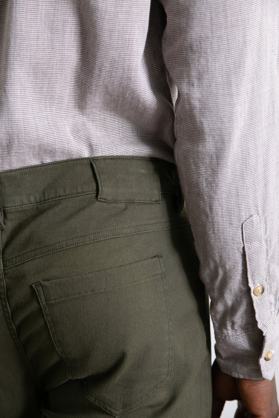 A close-up side view of a person wearing Bridge & Burn's Polk Pant in olive, featuring straight-fit organic cotton and 5-pocket styling, paired with a light long-sleeved shirt with buttoned cuffs. Only the torso and upper legs are shown.