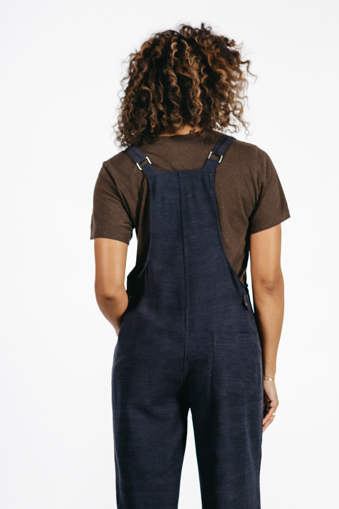 A person with curly hair, seen from behind, wears a brown t-shirt and Bridge & Burn's Edie Overall in Navy, featuring adjustable straps, against a plain white background.