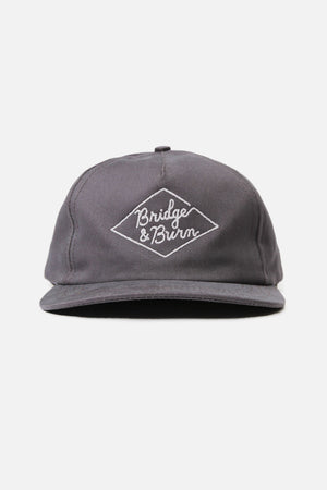 The Diamond Head Cap / Grey by Bridge & Burn is a gray cotton twill hat featuring “Bridge & Burn” embroidered in white inside a diamond shape on the front. It has a leather adjustment strap at the back, is made in the USA, and is shown against a plain white background.